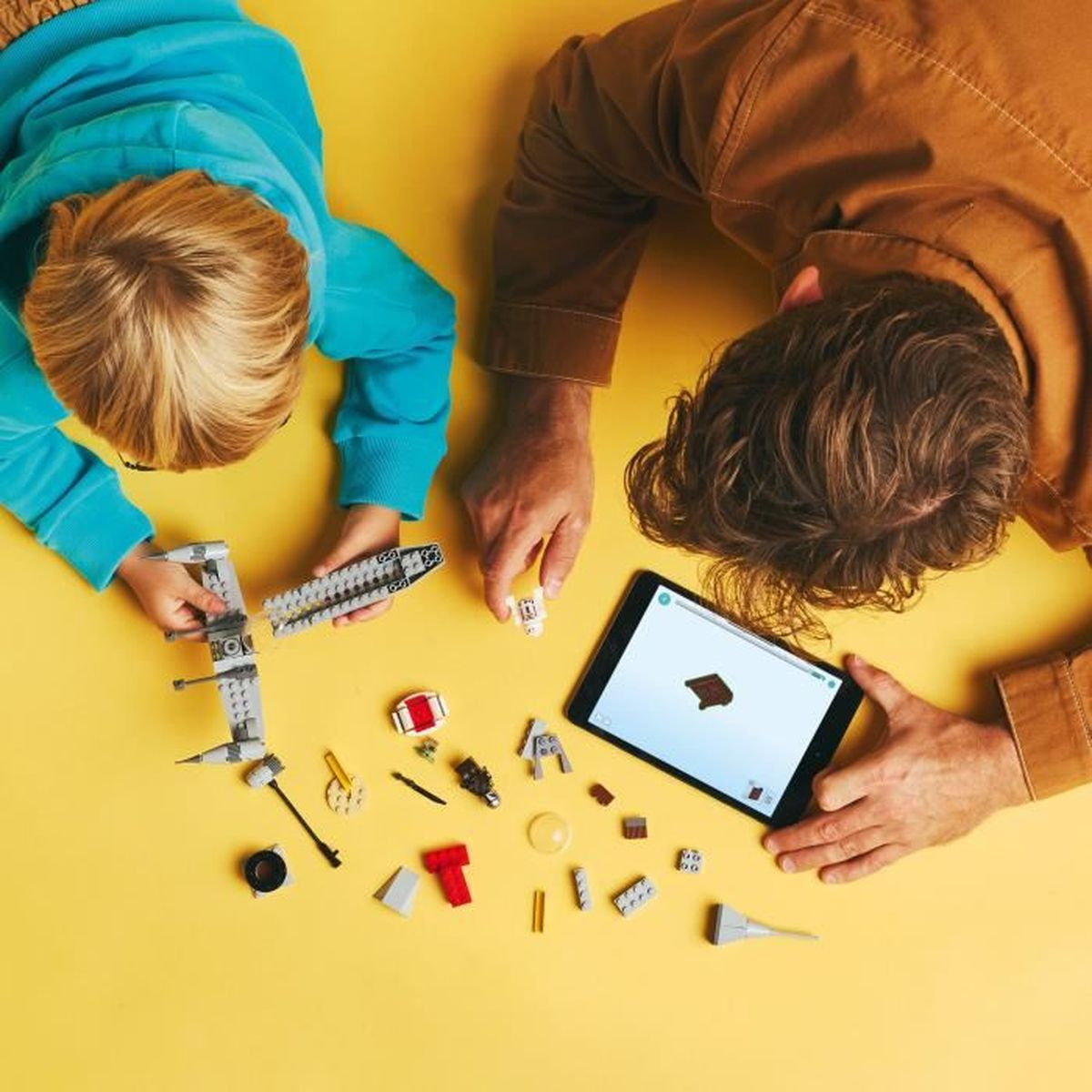 Dos personas montan un set de Lego sobre una superficie amarilla; instrucciones en una tableta.