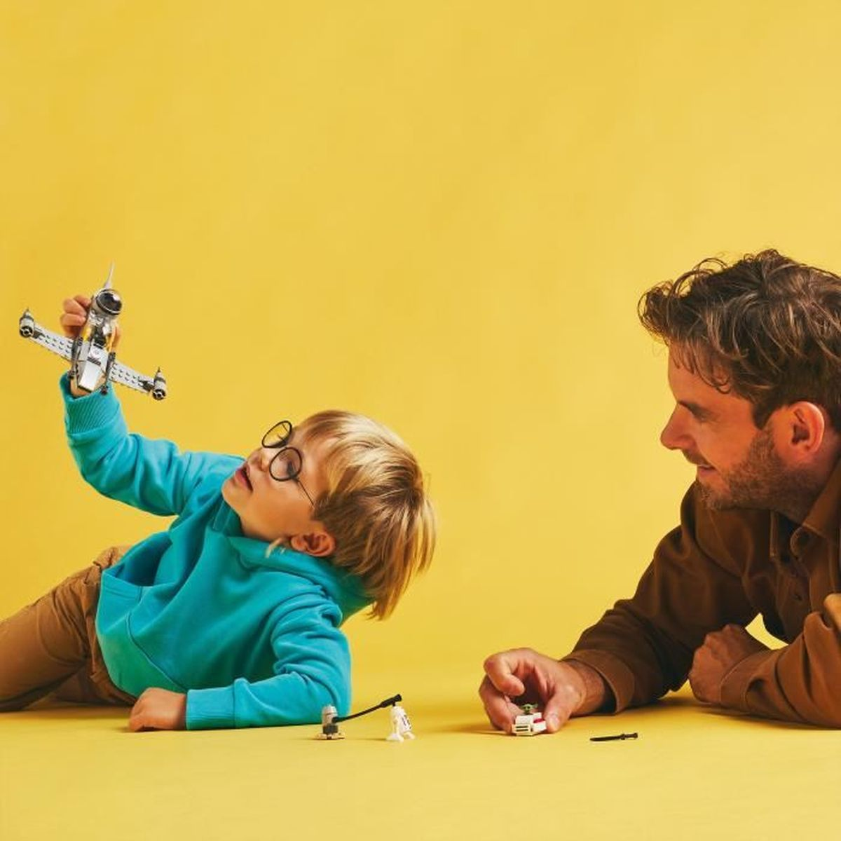 Un niño y un adulto juegan con un avión Lego, sobre un fondo amarillo.