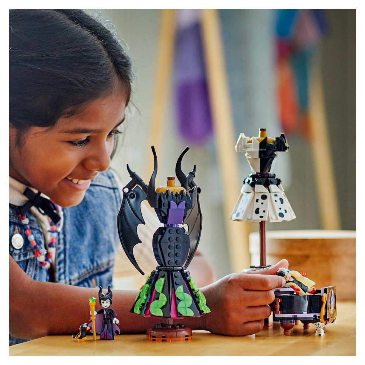 Dziewczynka uśmiecha się i bawi zestawem Lego z formami sukienek Maleficent i Cruelli oraz minifigurkami.