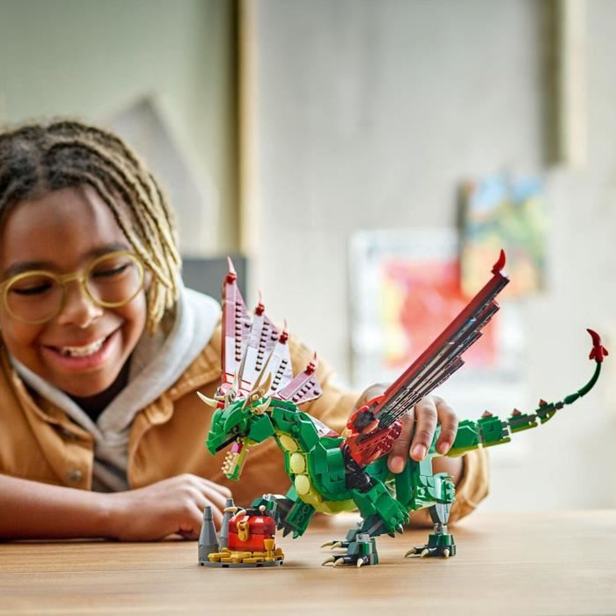 Dziecko uśmiecha się do zielonego smoka LEGO. Smok ma czerwone skrzydła obok skrzyni skarbów.