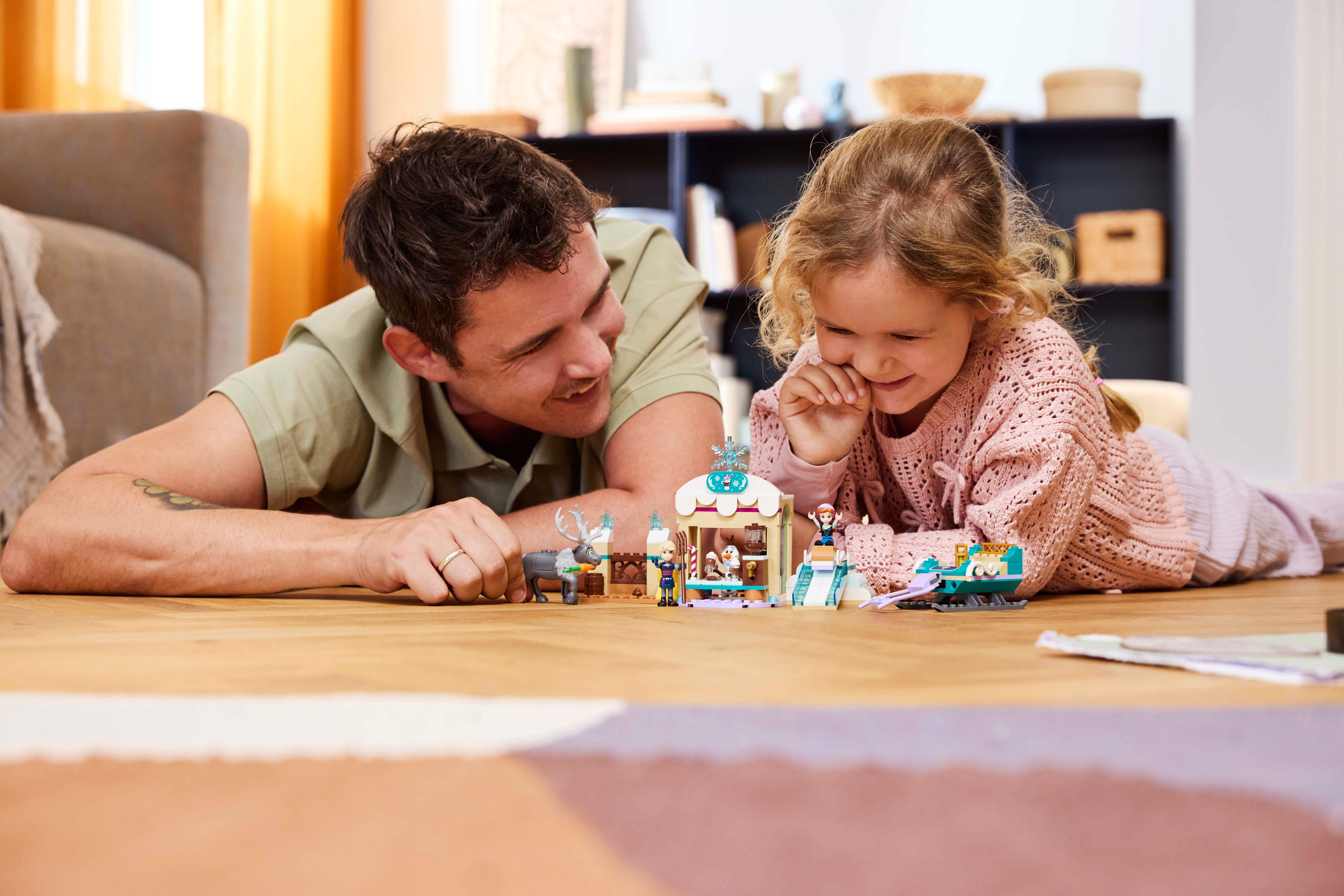 Un padre y una hija juegan con un set de Lego en un suelo de madera. El set incluye un edificio.