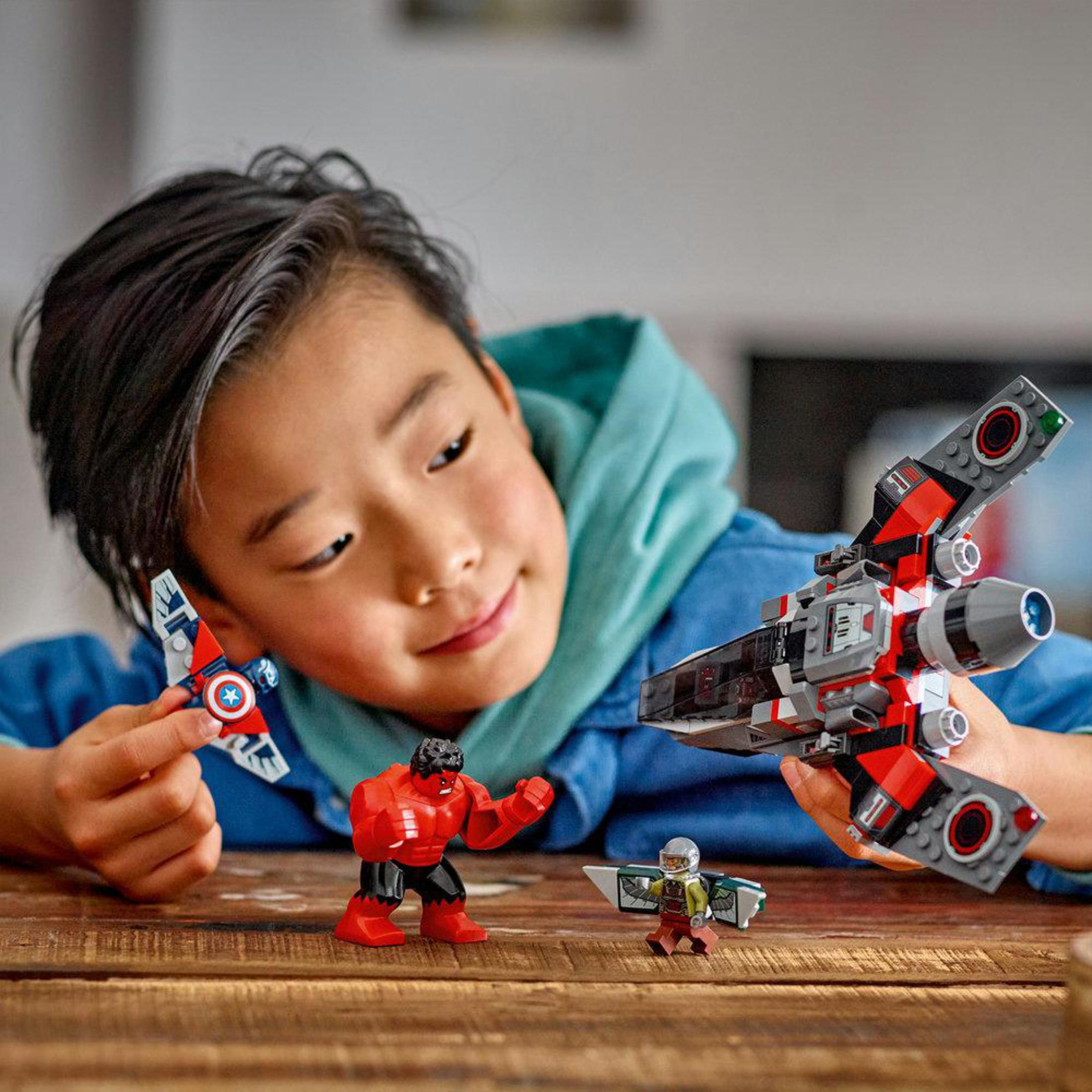 Junge mit LEGO-Spielzeug: Flugzeug, Hulk, Düsenjet. Holztisch, blauer Hemdhintergrund.