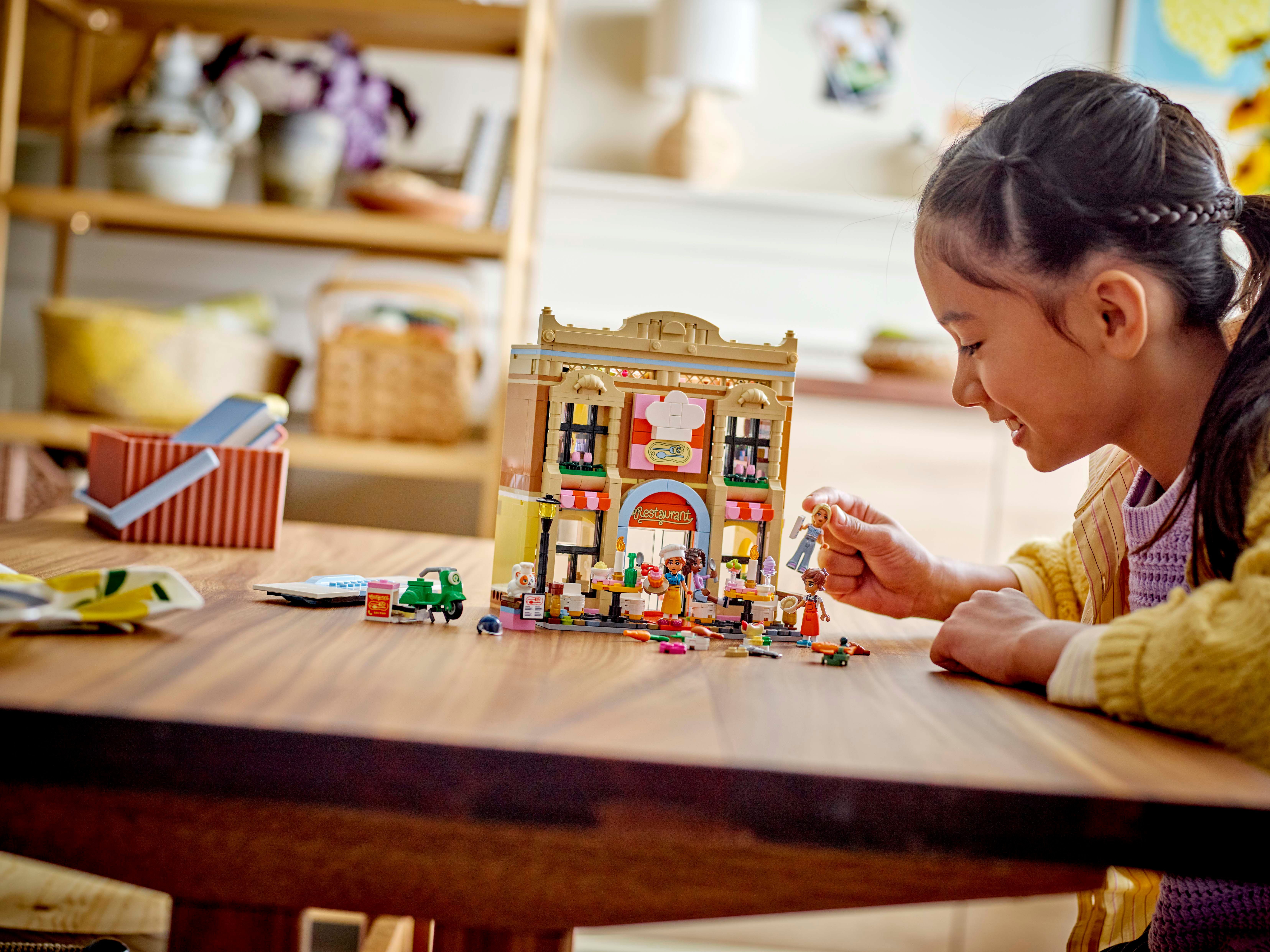 Dziewczynka bawi się restauracją Lego, drewniany stół. Tło: półki, akcesoria kuchenne.