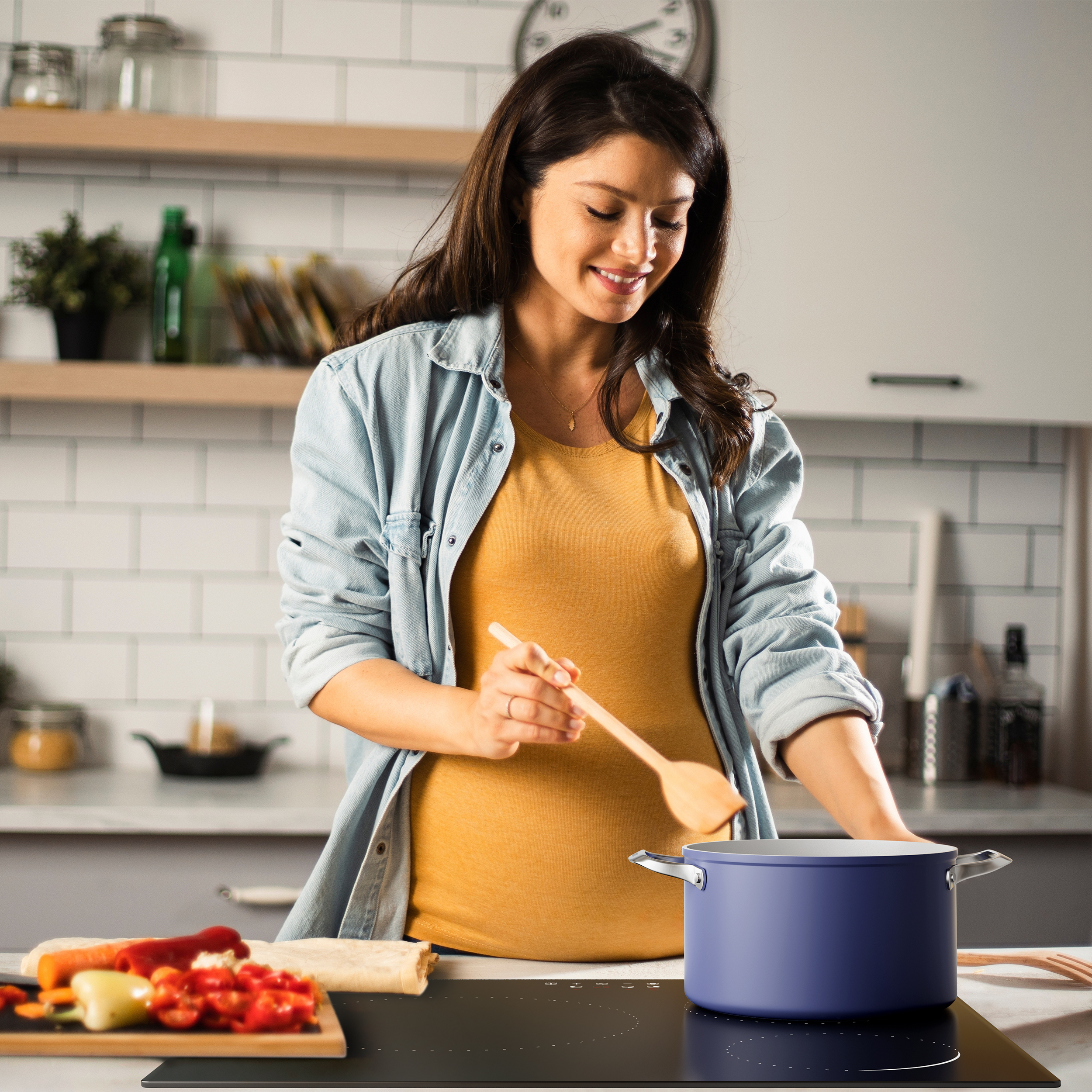 A woman in a kitchen stirring a pot on a black stove. Vegetables on a cutting board.