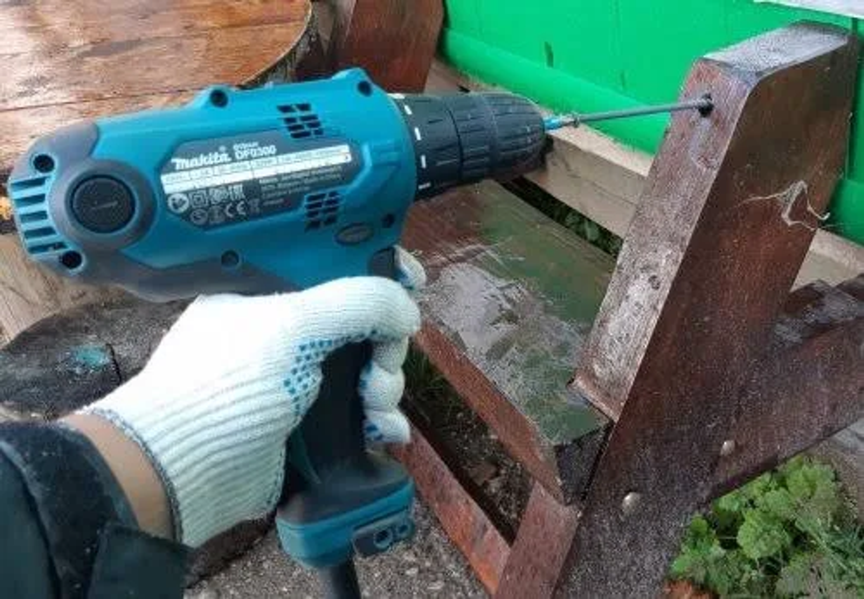 Osoba trzyma turkusową wiertarkę Makita ze śrubą wkręconą w drewnianą konstrukcję, w białej rękawicy.