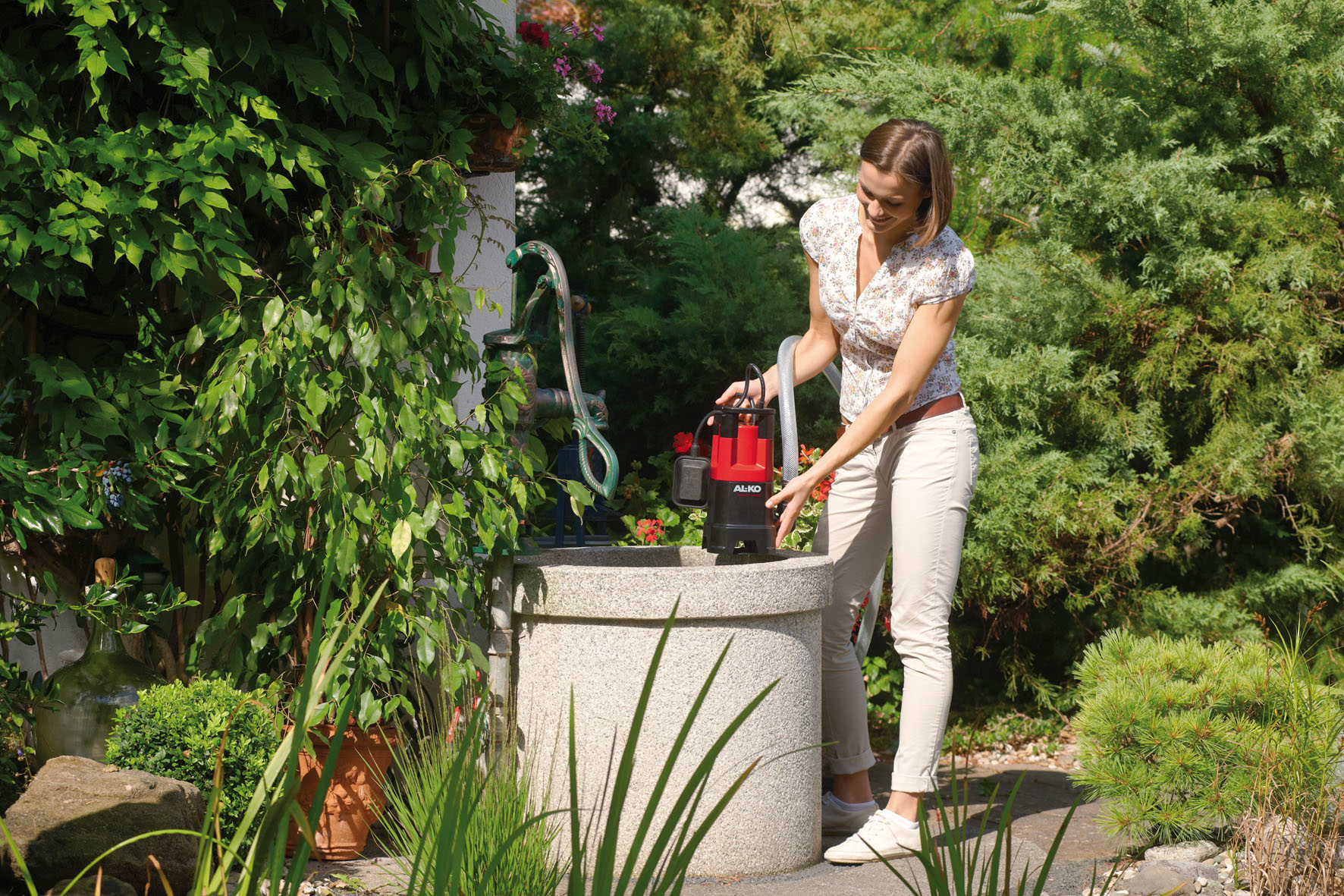 Une femme dans un jardin avec une pompe à eau. Elle tient une pompe noire et rouge près d'un puits.