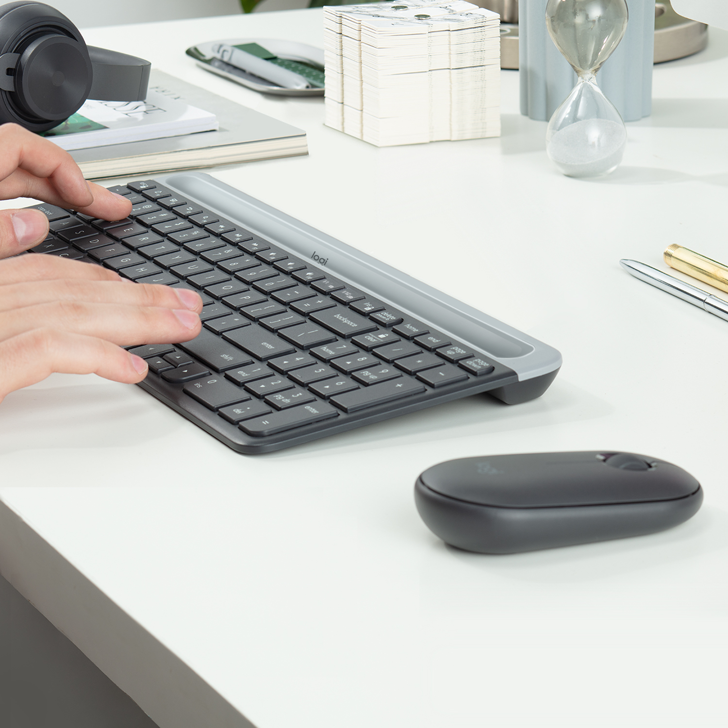 Manos escribiendo en un teclado gris con un ratón negro sobre un escritorio blanco. Hay auriculares.