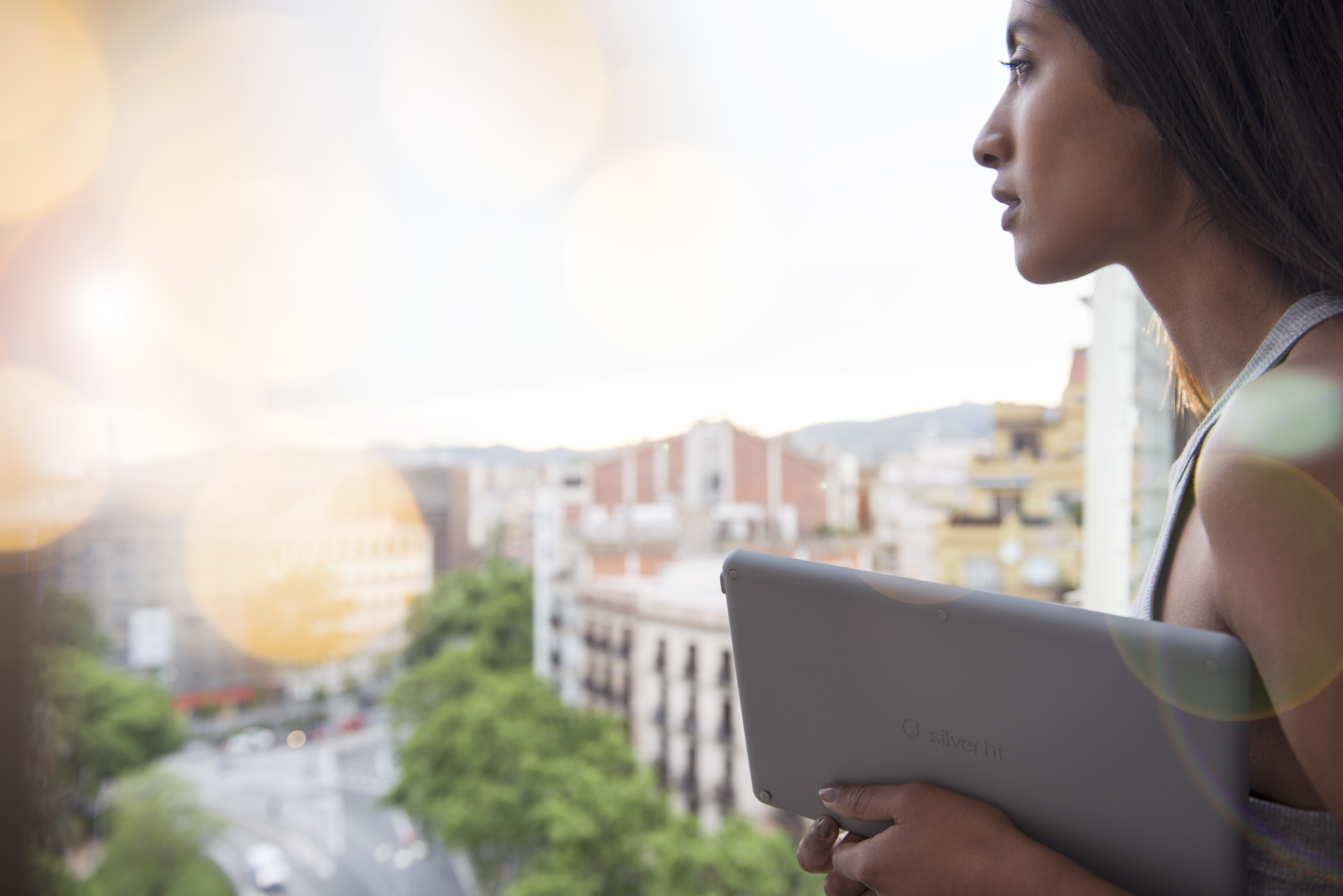 Kobieta wyglądająca przez okno, trzymająca tablet. W tle widoczne budynki miejskie i niebo.