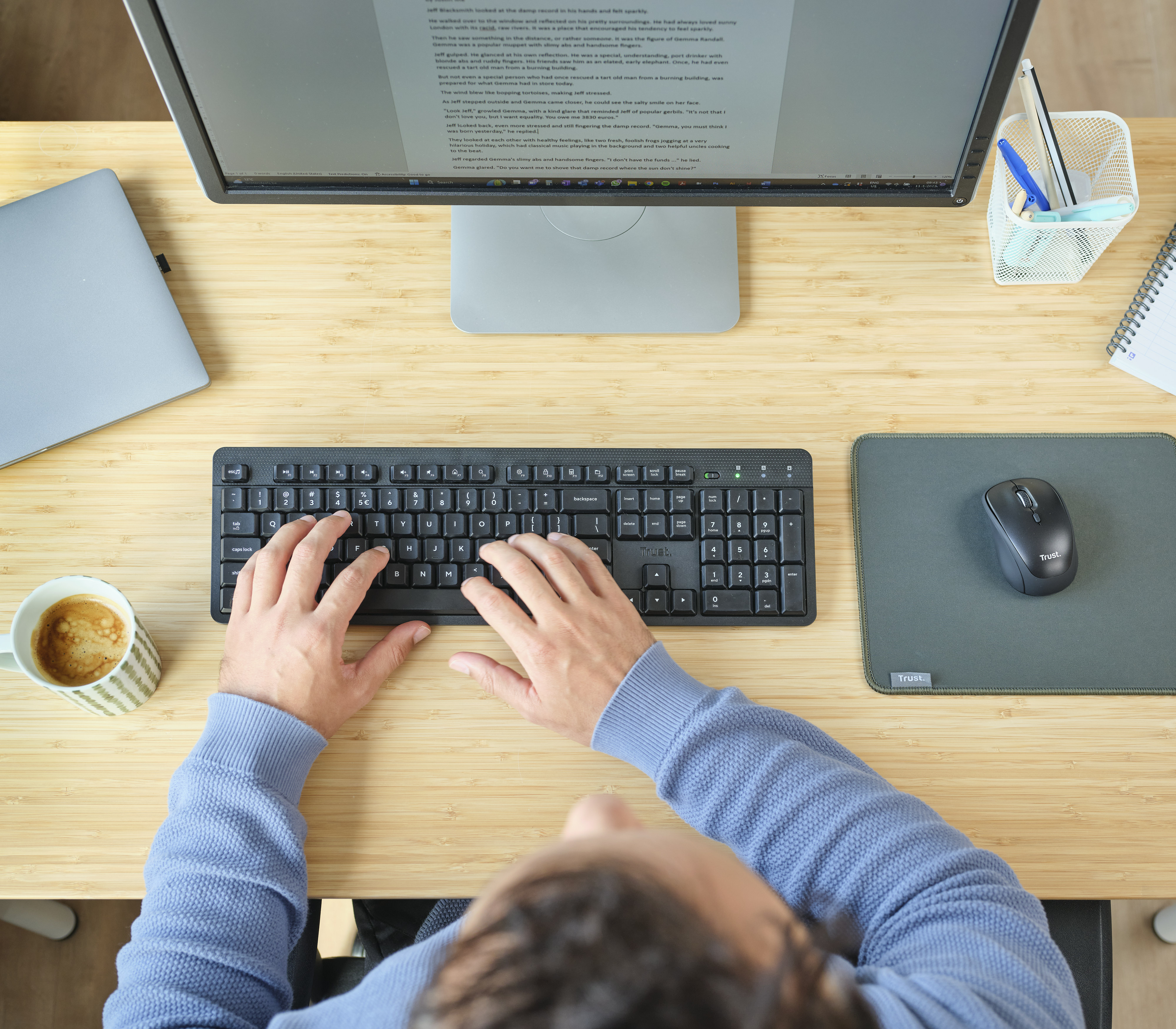 Osoba pisząca na czarnej klawiaturze. Jest monitor, mysz, kubek kawy i notatnik.