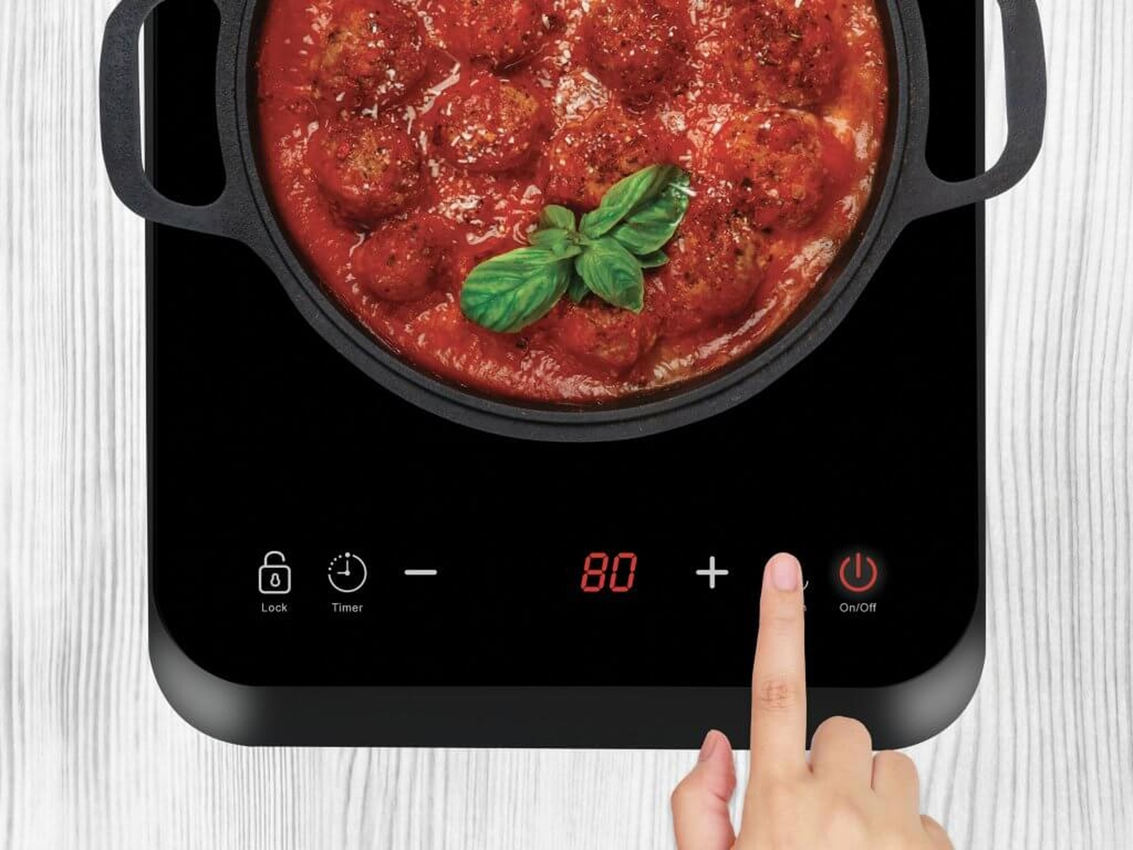 Zbliżenie garnka z klopsikami w sosie na płycie indukcyjnej, z ręką naciskającą przycisk.