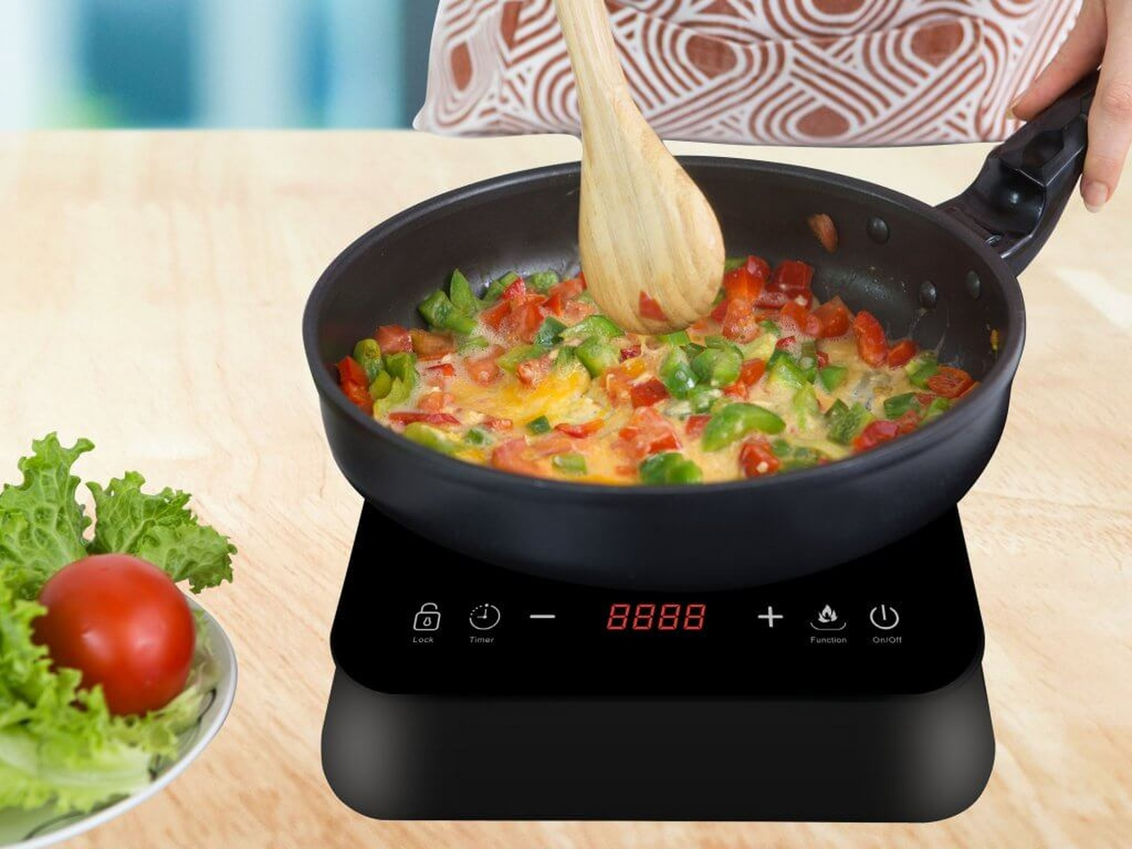 Patelnia na czarnej płycie indukcyjnej z gotującym się jedzeniem i drewnianą łyżką. W pobliżu mała sałatka i pomidor.