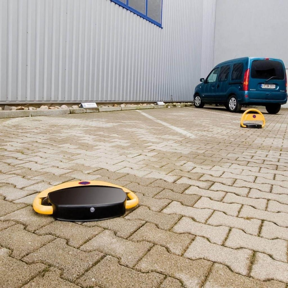 Parkplatz mit blauem Van. Zwei gelbe Parkplatzschützer auf Backsteinpflaster.