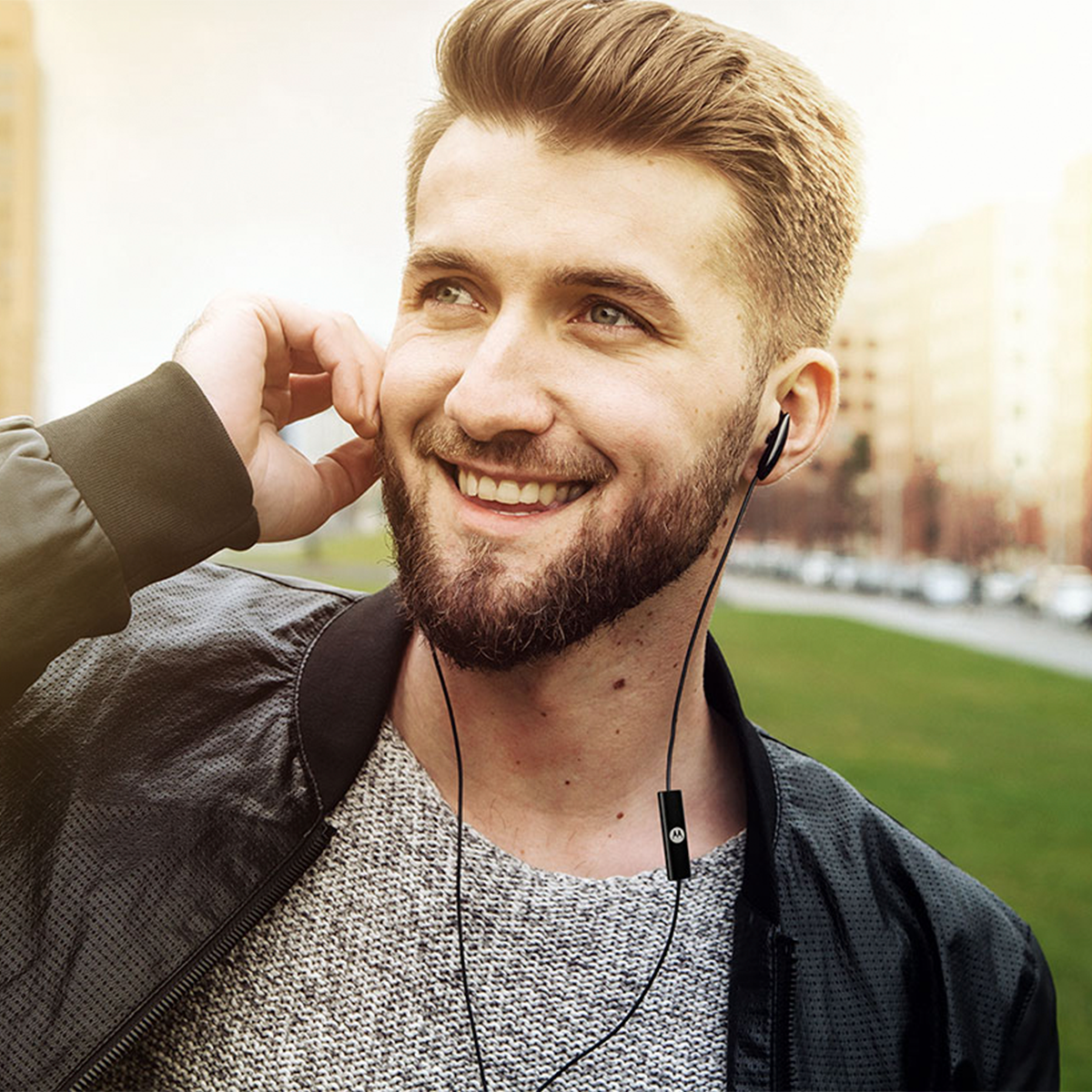 Mężczyzna z brodą w szarym swetrze i czarnych słuchawkach, uśmiechnięty.