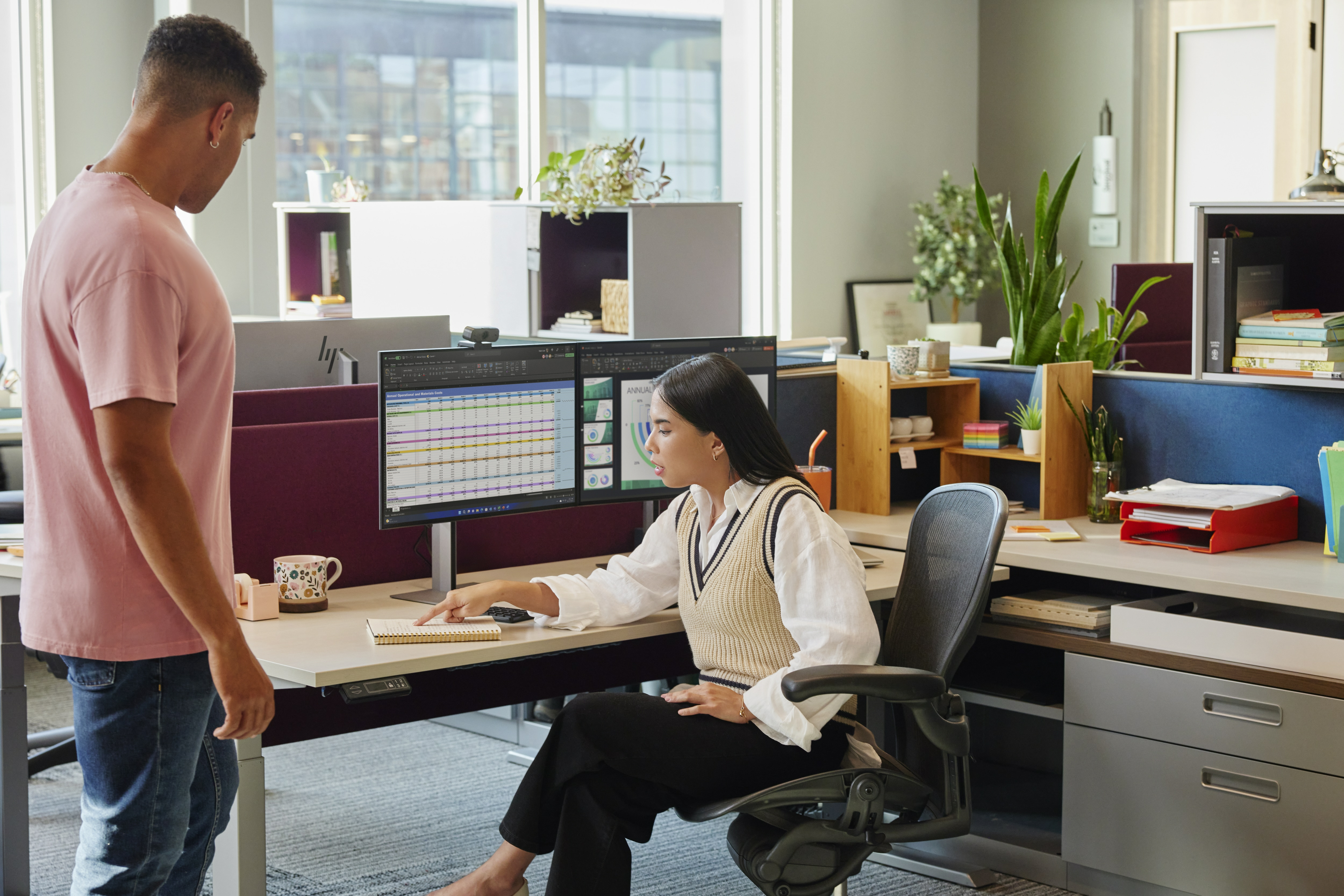 Dwie osoby w biurze, patrzące na podwójne monitory komputerowe. Scena z nowoczesną konfiguracją.