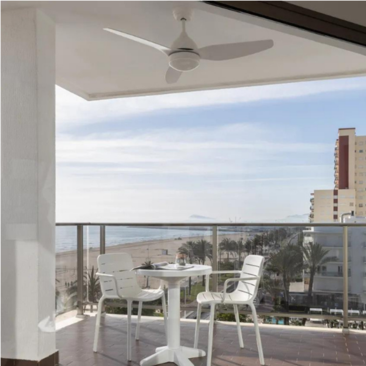 Vista dal balcone con un ventilatore da soffitto bianco. Tavoli, sedie, mare, spiaggia ed edifici in lontananza.