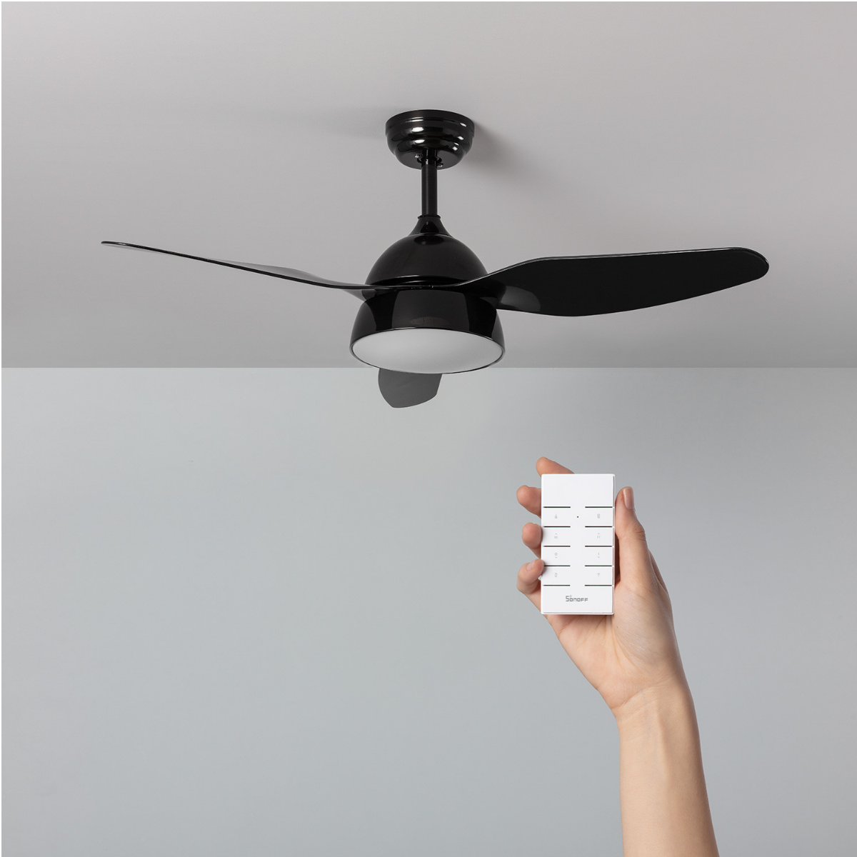 Ventilatore a soffitto nero con telecomando in mano, su sfondo grigio.