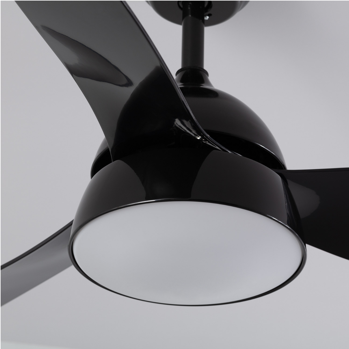 Primo piano di un ventilatore da soffitto nero con tre pale lucide e un'apparecchio di illuminazione bianco. Il ventilatore è contro un soffitto chiaro.
