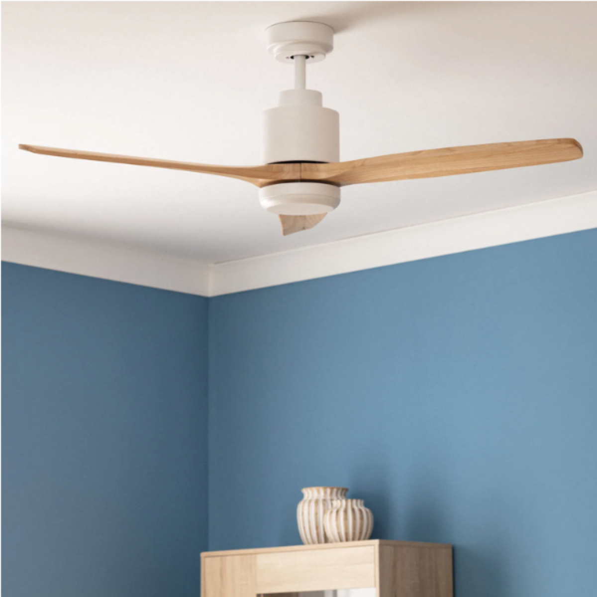 Ventilatore da soffitto bianco con pale in legno in una stanza con parete blu. Ci sono due vasi su un mobile.