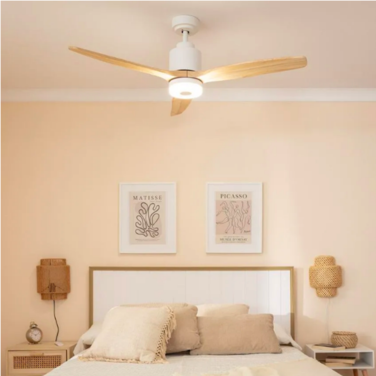 Un ventilatore da soffitto bianco con pale in legno è sopra un letto. Due opere d'arte sul muro.