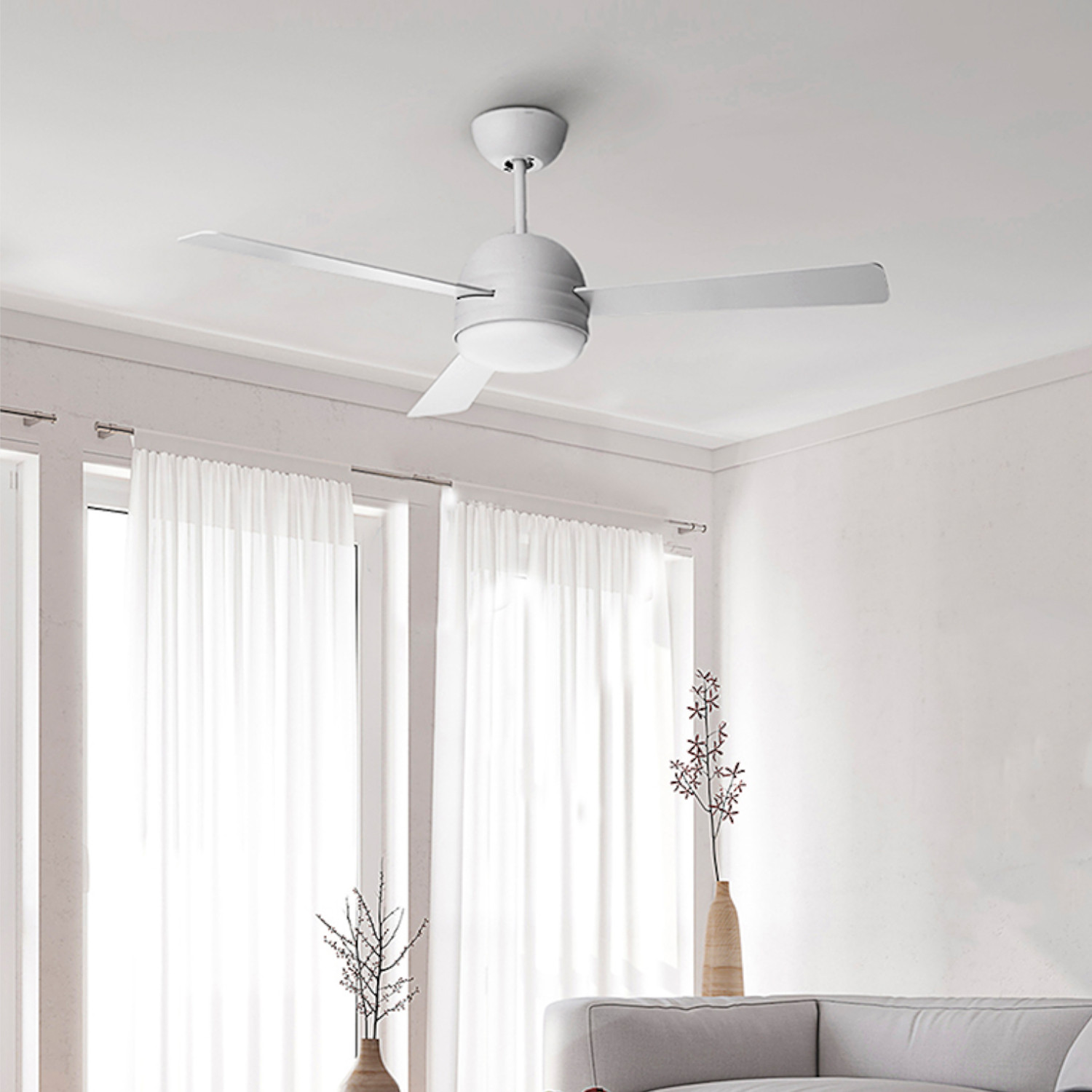 Una stanza con ventilatore a soffitto, tende trasparenti e vasi con piante. Un divano grigio in primo piano.