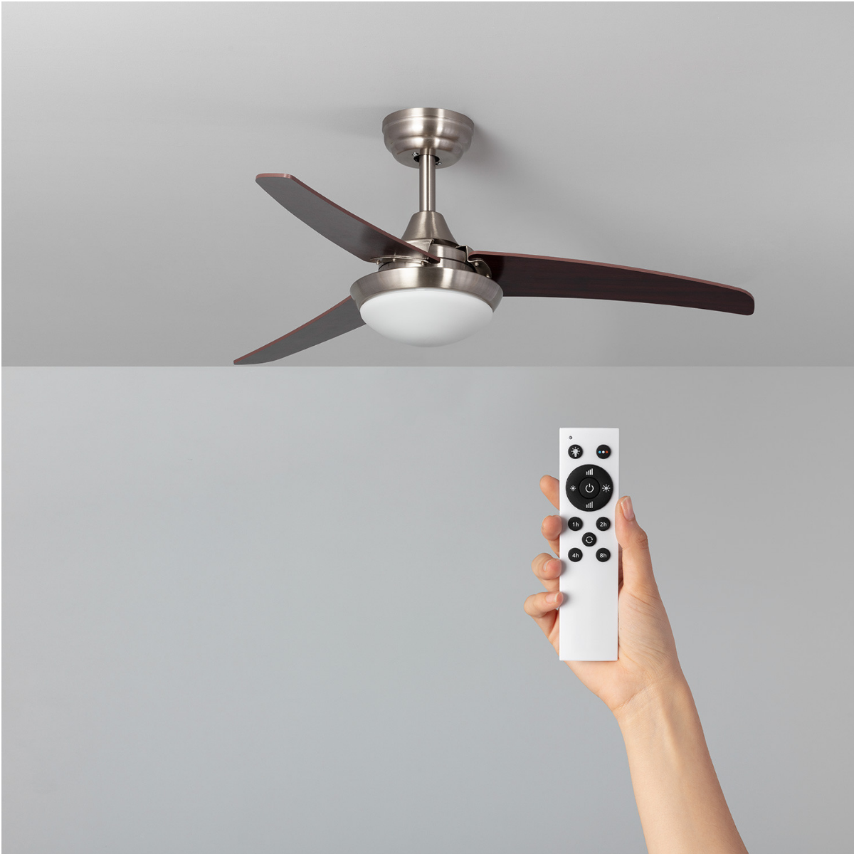 Ventilatore da soffitto con tre pale marrone scuro, una luce e un telecomando in mano, su sfondo grigio.