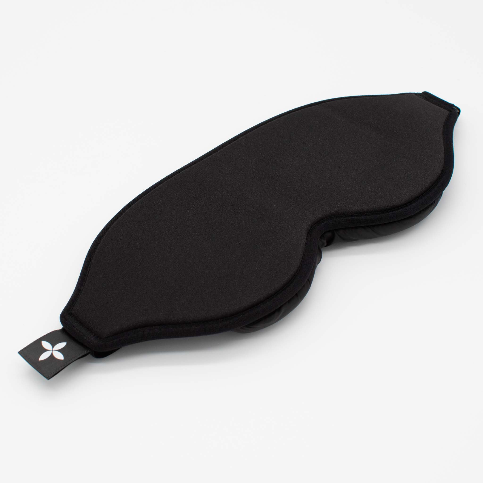 Eine schwarze Schlafmaske, konturierte Form und ein weißes Logo-Etikett. Weißer Hintergrund.