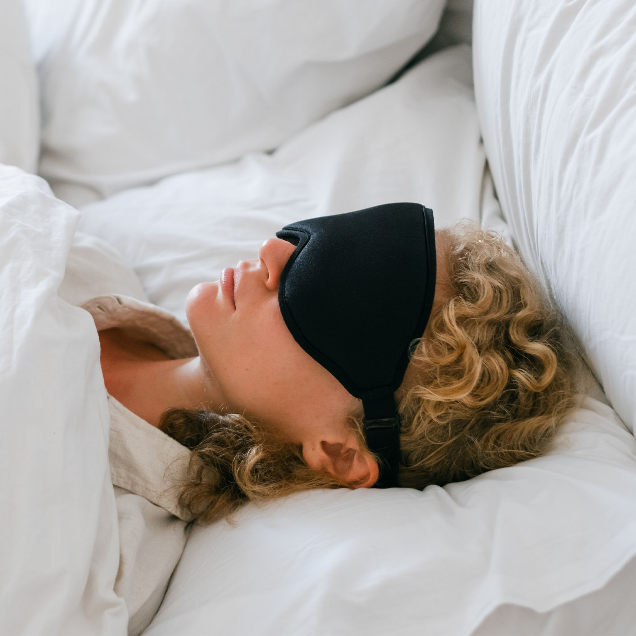 Eine Person mit schwarzer Schlafmaske liegt auf einem weißen Bett, Gesicht und Haare sichtbar.