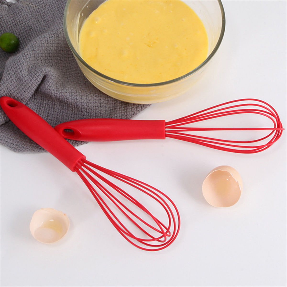 Zwei rote Schneebesen und eine Schüssel mit gelbem Teig, mit Eierschalen auf weißer Oberfläche.