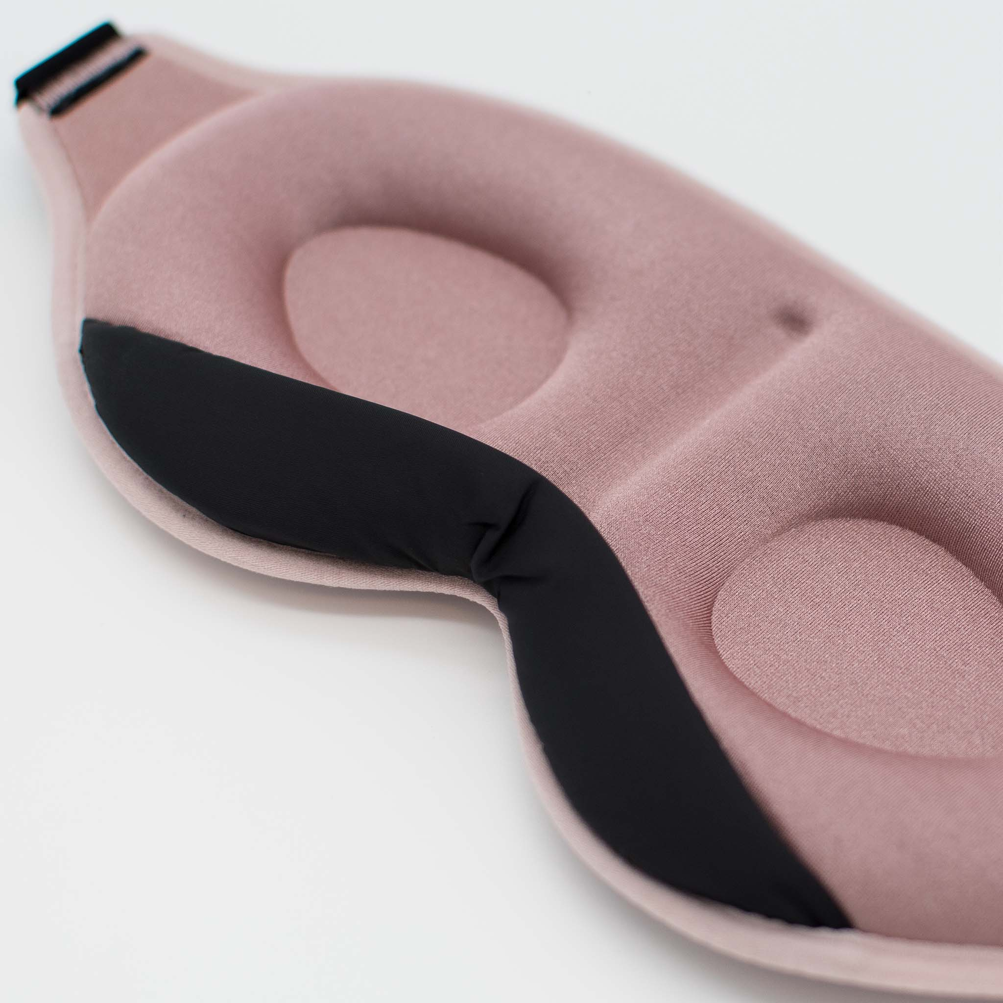 Eine rosa Schlafmaske liegt auf einer weißen Oberfläche. Sie hat eine gebogene Form mit einem schwarzen Nasenstück.