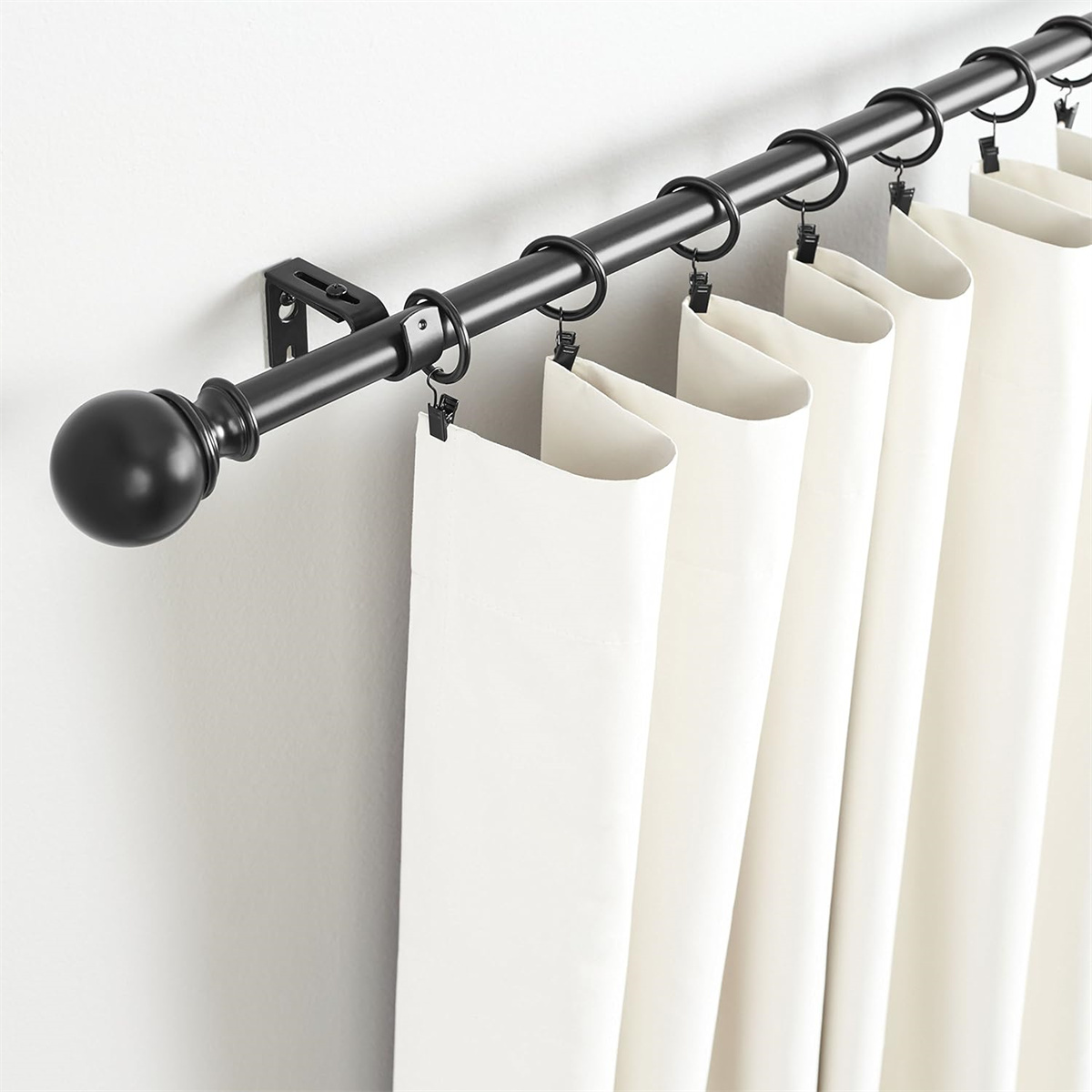 Schwarze Gardinenstange mit Ringen und weißen Vorhängen. Stange ist an einer weißen Wand befestigt.