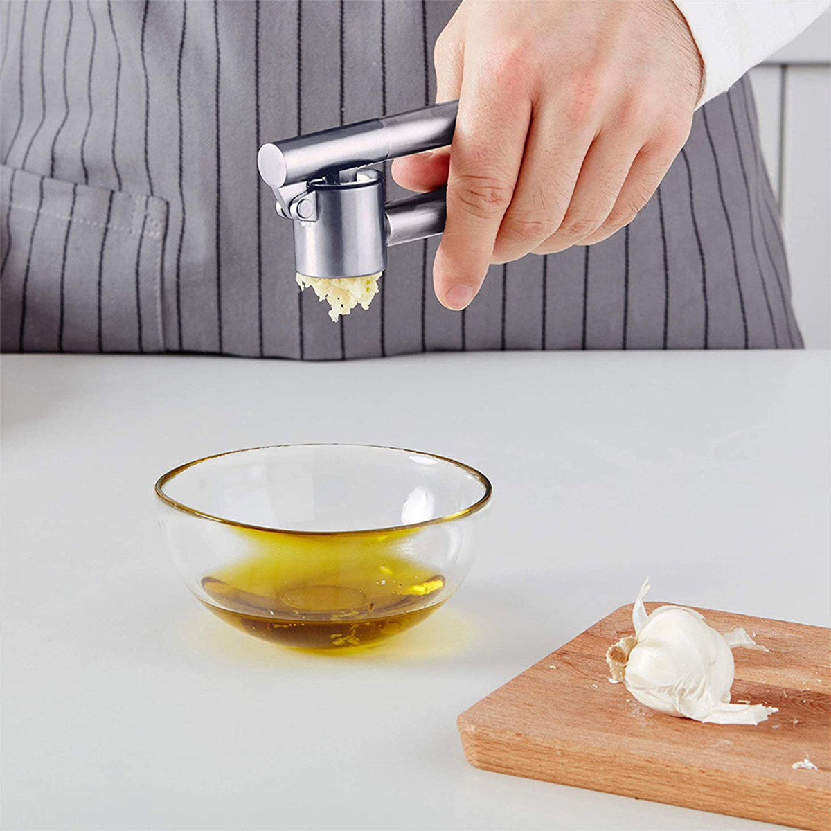 Eine Hand drückt eine Knoblauchpresse. Gehackter Knoblauch fällt in eine Schüssel. Olivenöl und eine Knoblauchzehe sind sichtbar.