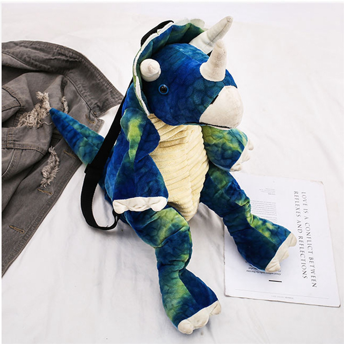 Ein blau-grüner Dinosaurier-Rucksack liegt auf einem Bett, im Hintergrund eine Jeansjacke.
