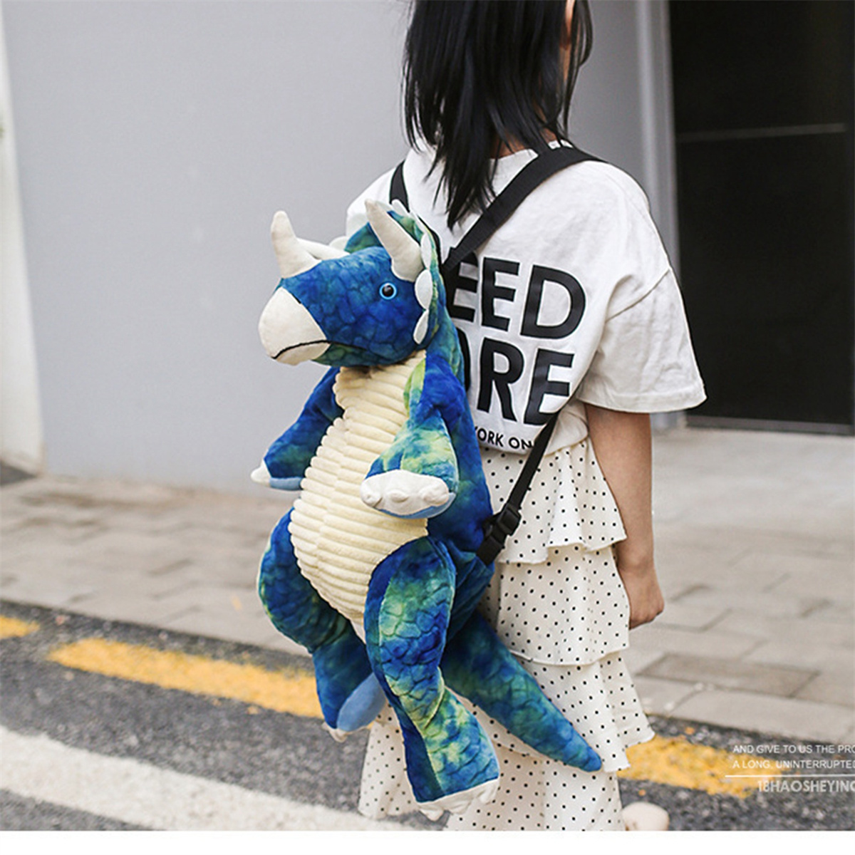 Ein Kind mit einem Dinosaurier-Rucksack. Der Rucksack ist blau und grün. Das Kind ist auf grauem Pflaster.