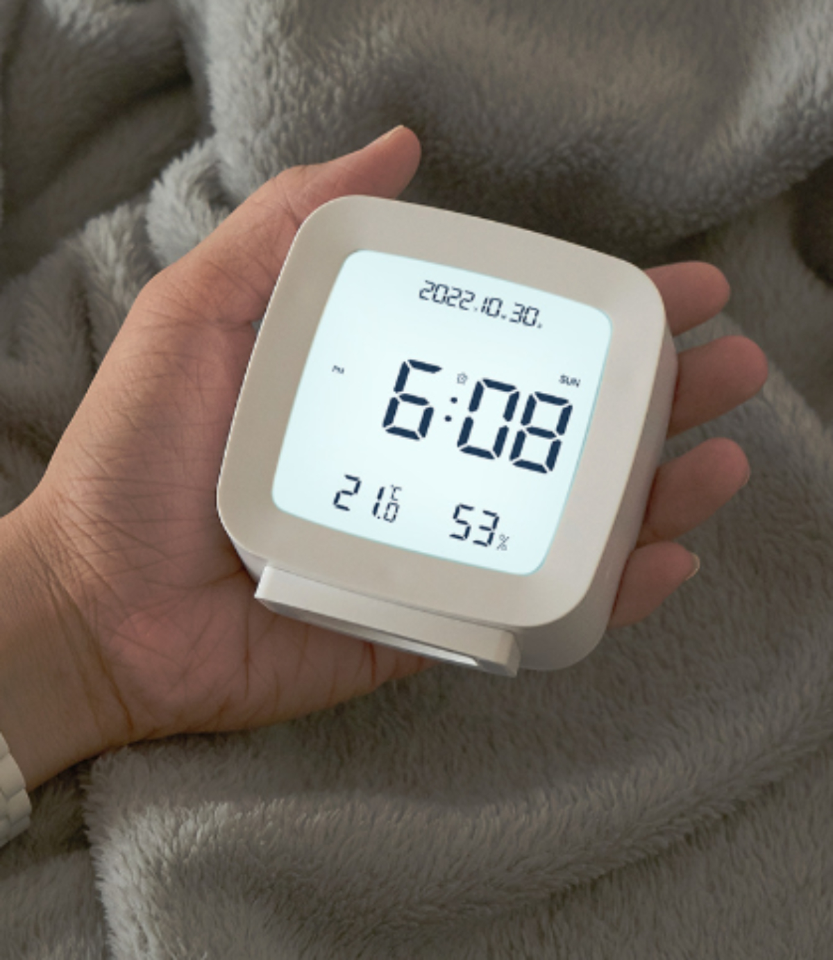 Ein weißer digitaler Wecker in einer Hand, der Datum, Uhrzeit, Temperatur und Luftfeuchtigkeit anzeigt.