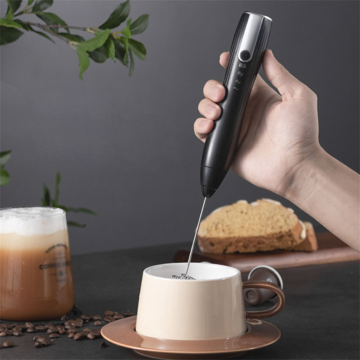 Eine Hand hält einen schwarzen Milchaufschäumer über eine beige Tasse. Ein Glas Kaffee und ein Stück Brot stehen auf dem Tisch.