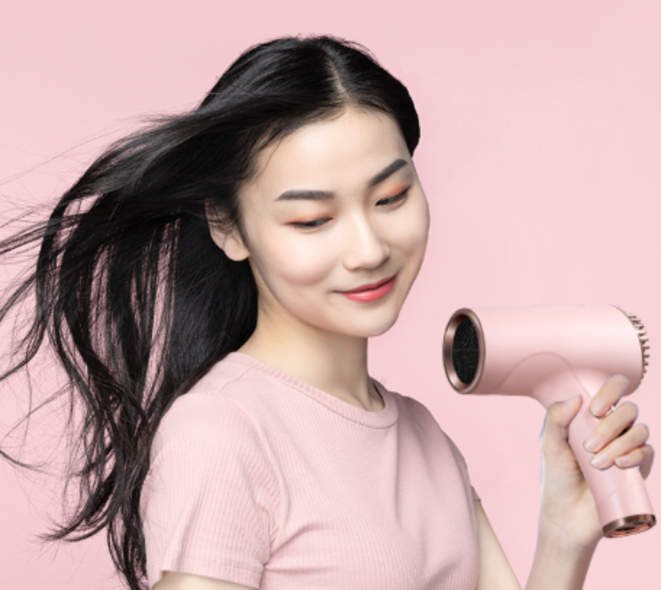 Eine Frau mit langen schwarzen Haaren hält einen rosa Haartrockner und lächelt vor rosa Hintergrund.