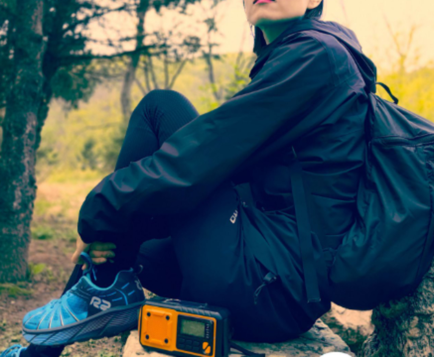 Person in schwarzer Kleidung sitzt mit Rucksack, Radio und blauem Schuh auf einem Felsen.