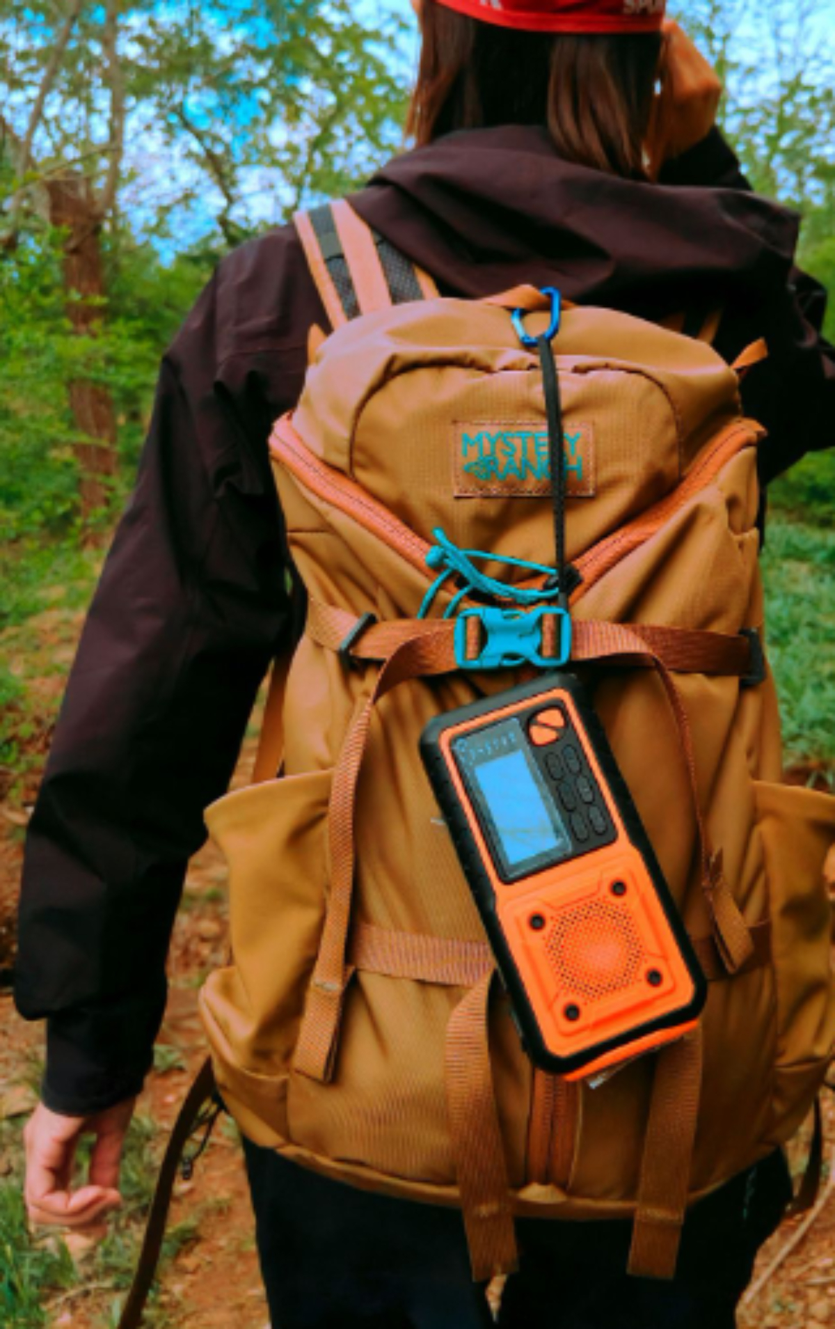Person mit Rucksack in einem Wald. Der Rucksack enthält ein Gerät mit orangefarbenem Bildschirm.