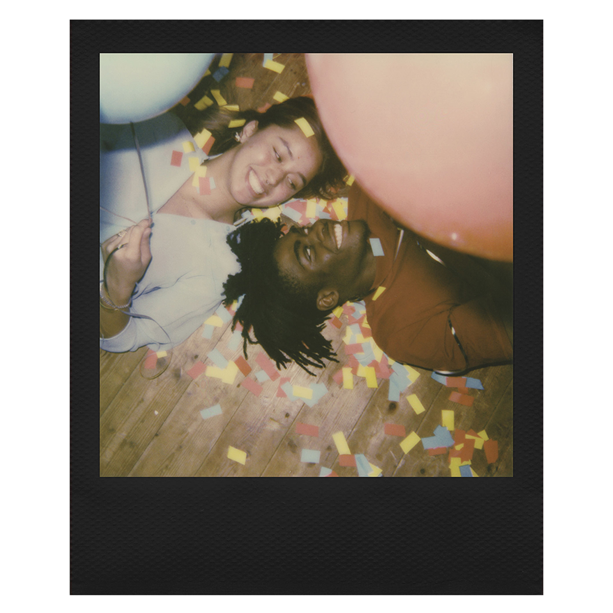 Zdjęcie polaroidowe uśmiechniętej pary leżącej na drewnianej podłodze z konfetti i dwoma balonami.