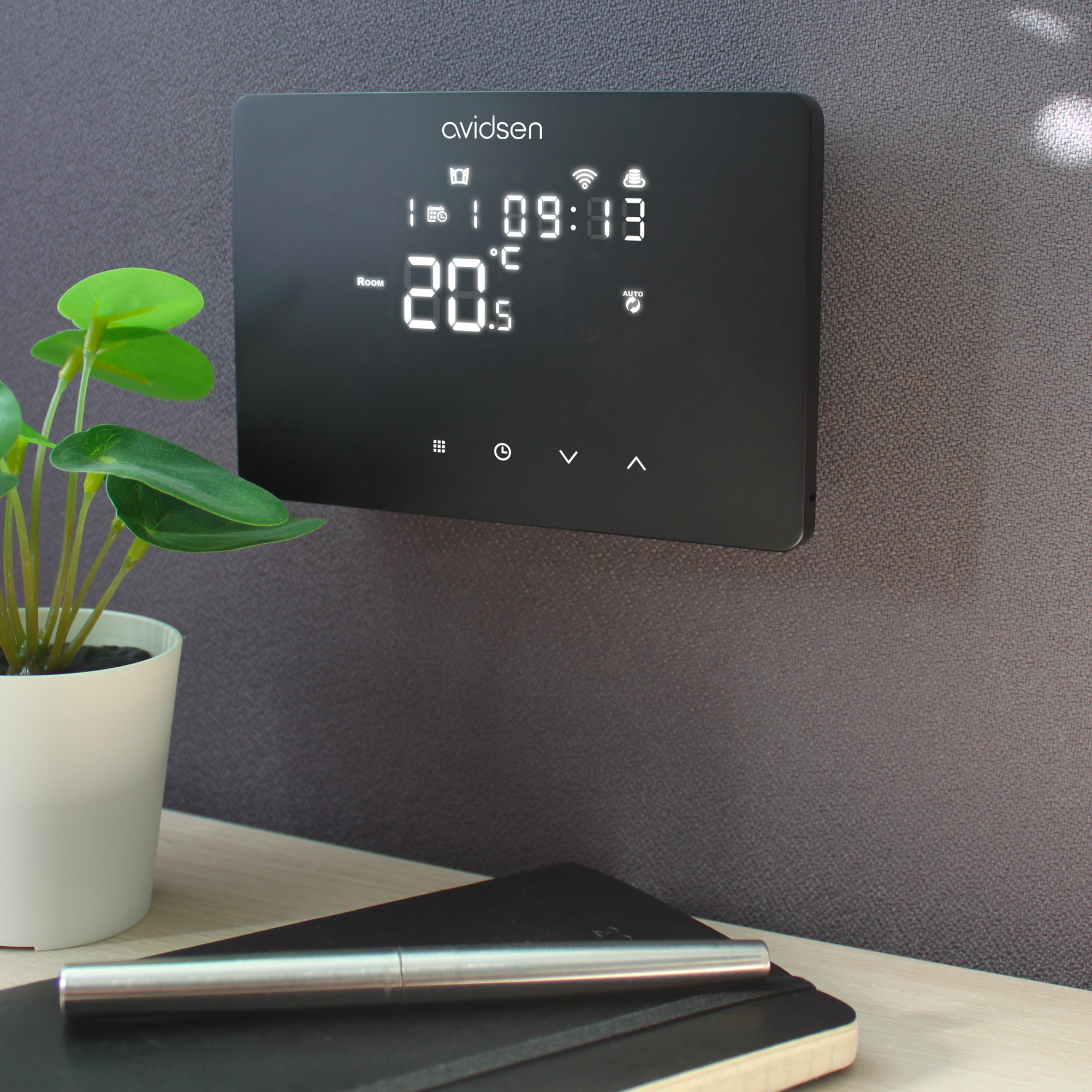 Termostato negro en pared gris que muestra hora, temperatura y ajustes. Planta y bolígrafo.