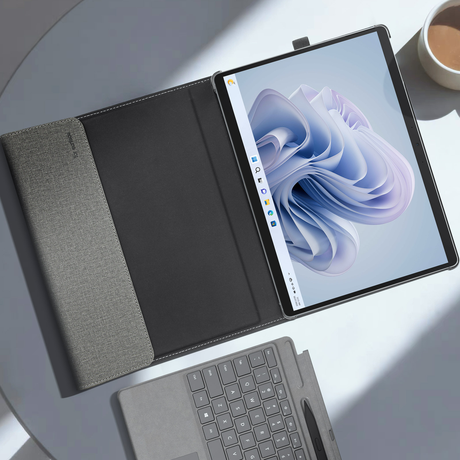 Tablet in custodia grigia, aperto su tavolo bianco. Tastiera e tazza di caffè visibili.