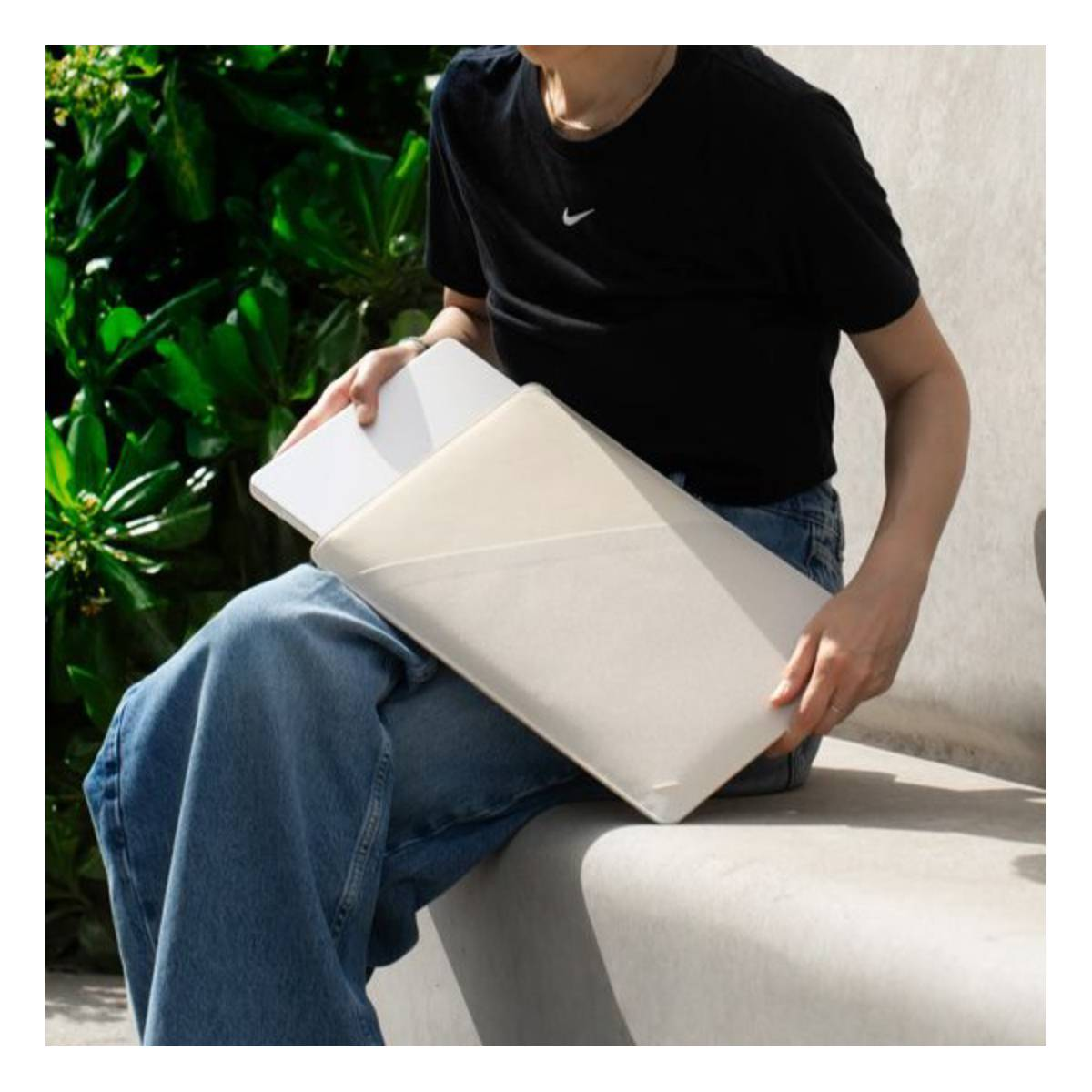 Personne assise avec étui pour ordinateur portable. Chemise noire, jean bleu. Feuillage vert en arrière-plan.