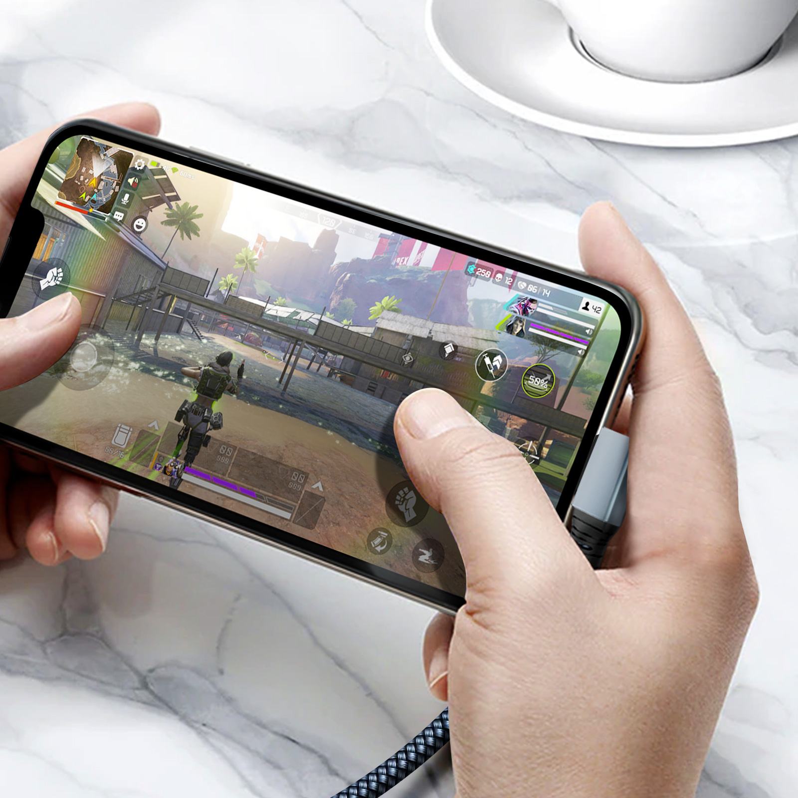 Mani che tengono un telefono che mostra un gioco. Tazza bianca e cavo visibili.