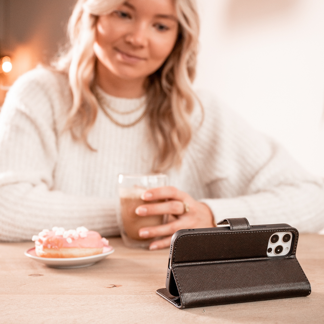 Femme tenant un verre de café au lait et regardant un téléphone dans un étui en cuir marron. Un beignet est sur la table.