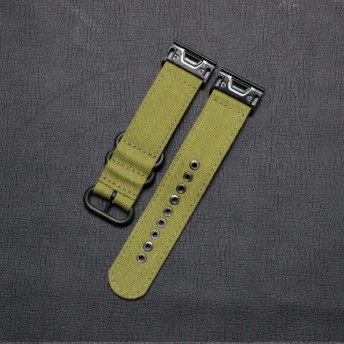 Zwei olivgrüne Uhrenarmbänder mit schwarzer Hardware, auf einer strukturierten, dunklen Oberfläche liegend.