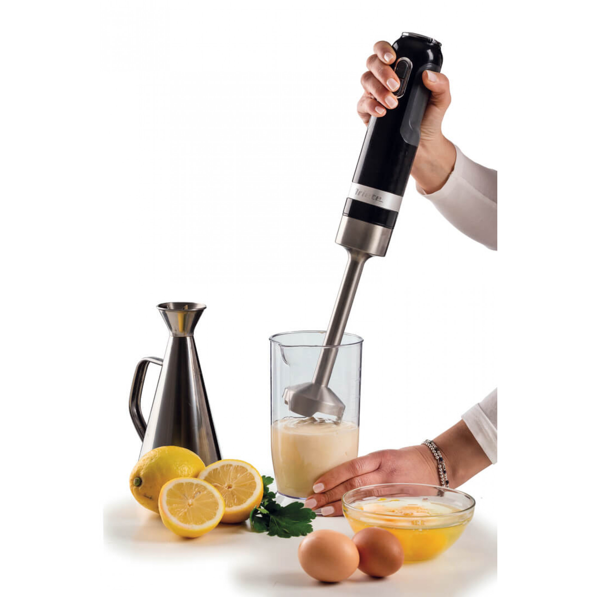 Osoba używająca czarnego i srebrnego blendera ręcznego. Składniki: cytryny, jajka, olej, pietruszka.