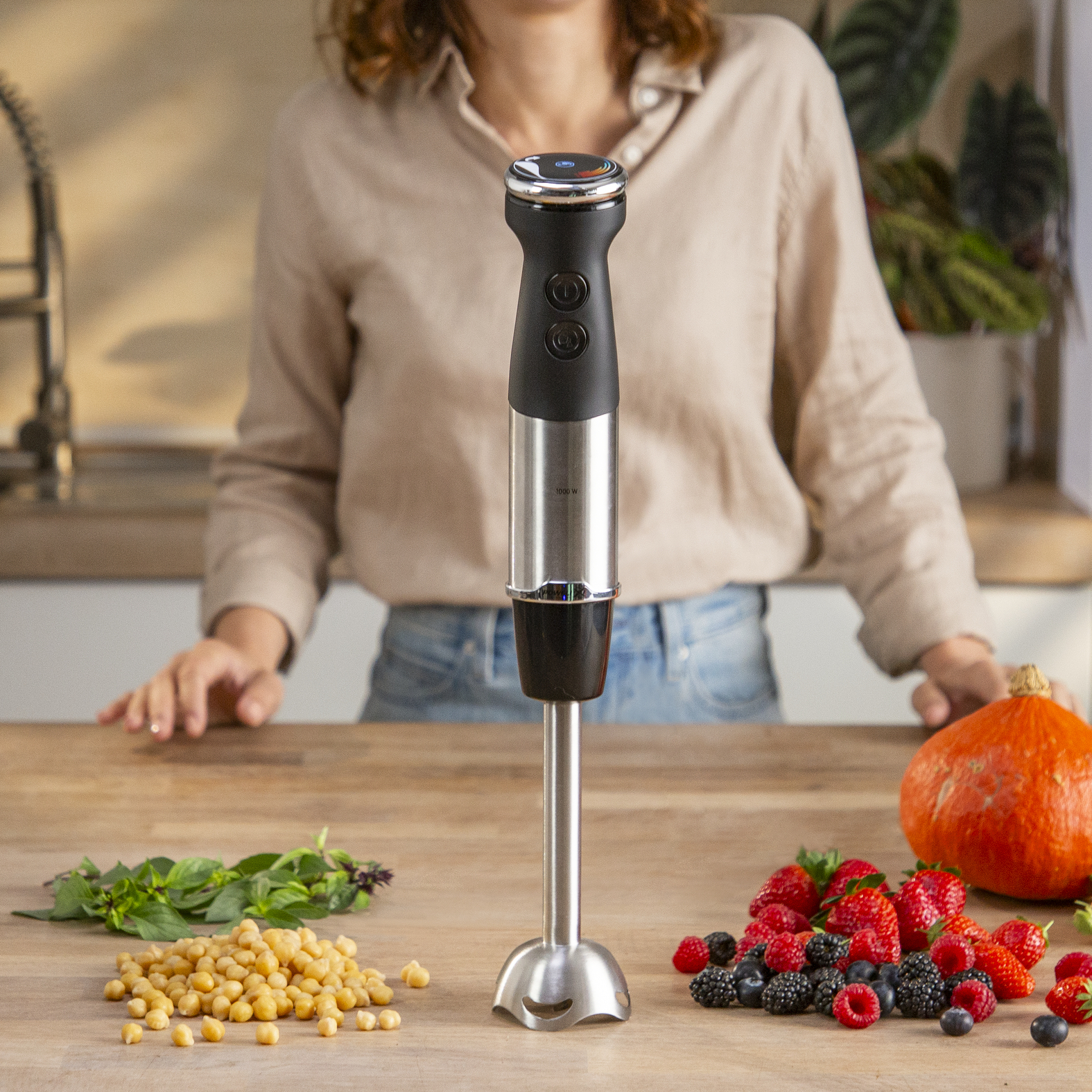 Kobieta używa blendera ręcznego. Warzywa i owoce na drewnianym stole.