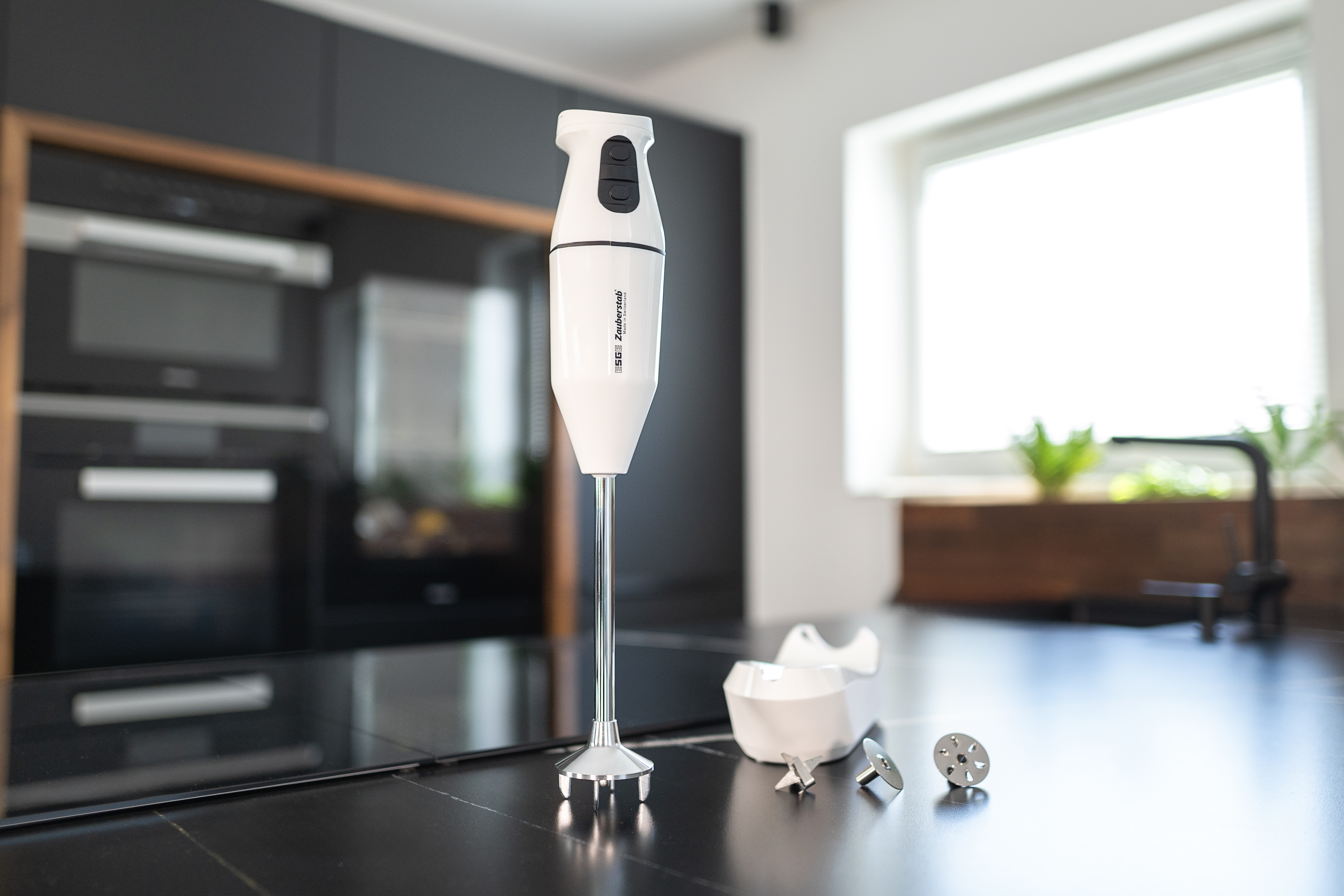 Ein weißer Handmixer mit Aufsätzen auf einer schwarzen Arbeitsplatte. Hintergrund: schwarze Schränke.