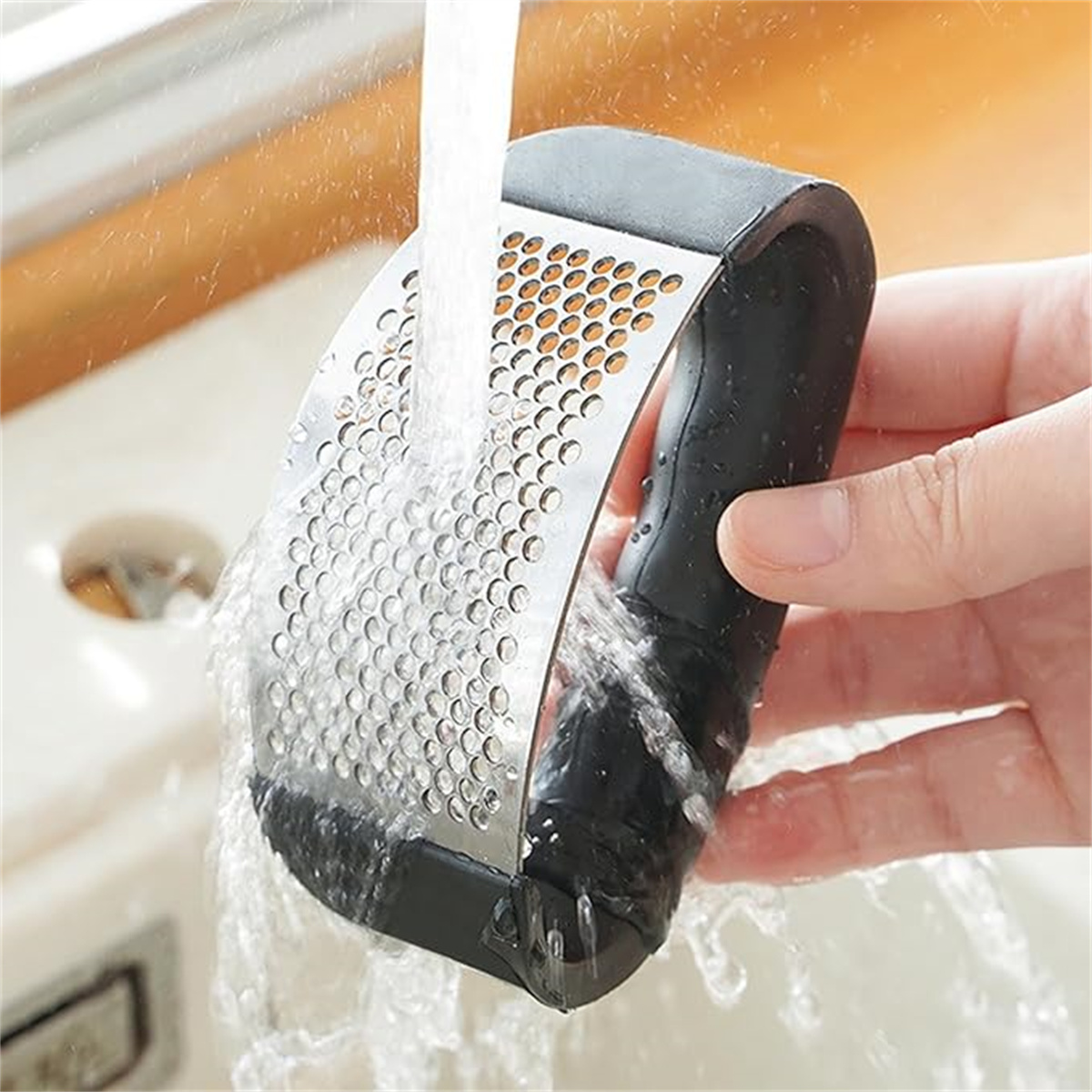 Hand einer Person, die eine Knoblauchpresse unter fließendem Wasser wäscht, Nahaufnahme auf hellfarbiger Oberfläche.