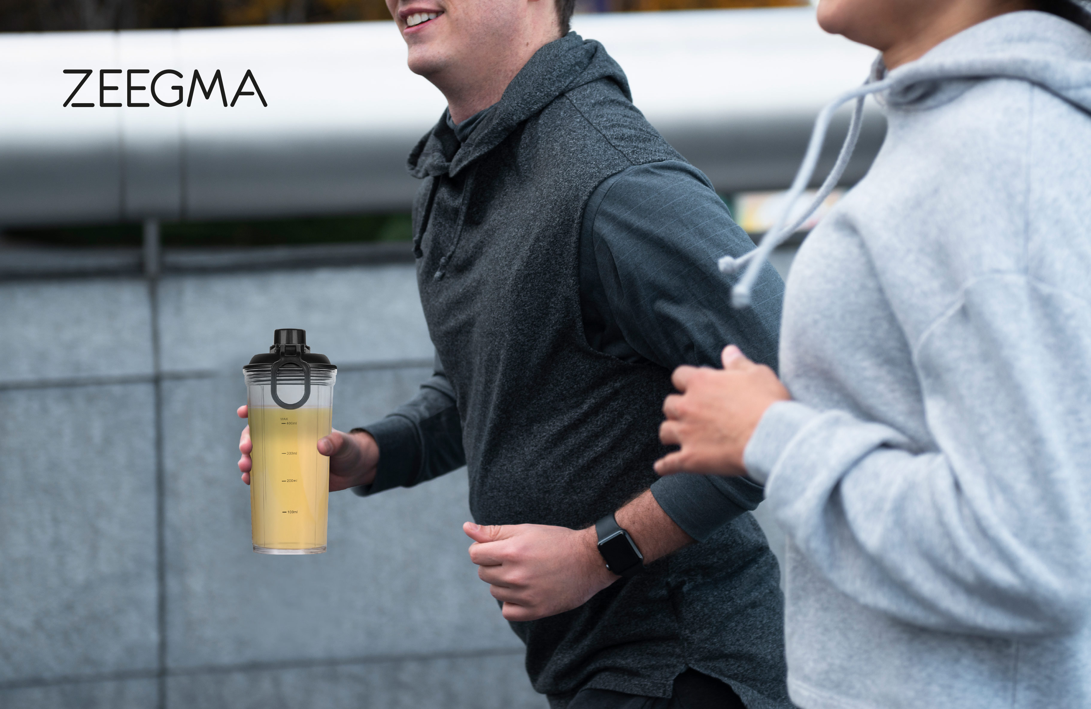 Dwie osoby biegną trzymając shaker z żółtym płynem, szare tło.